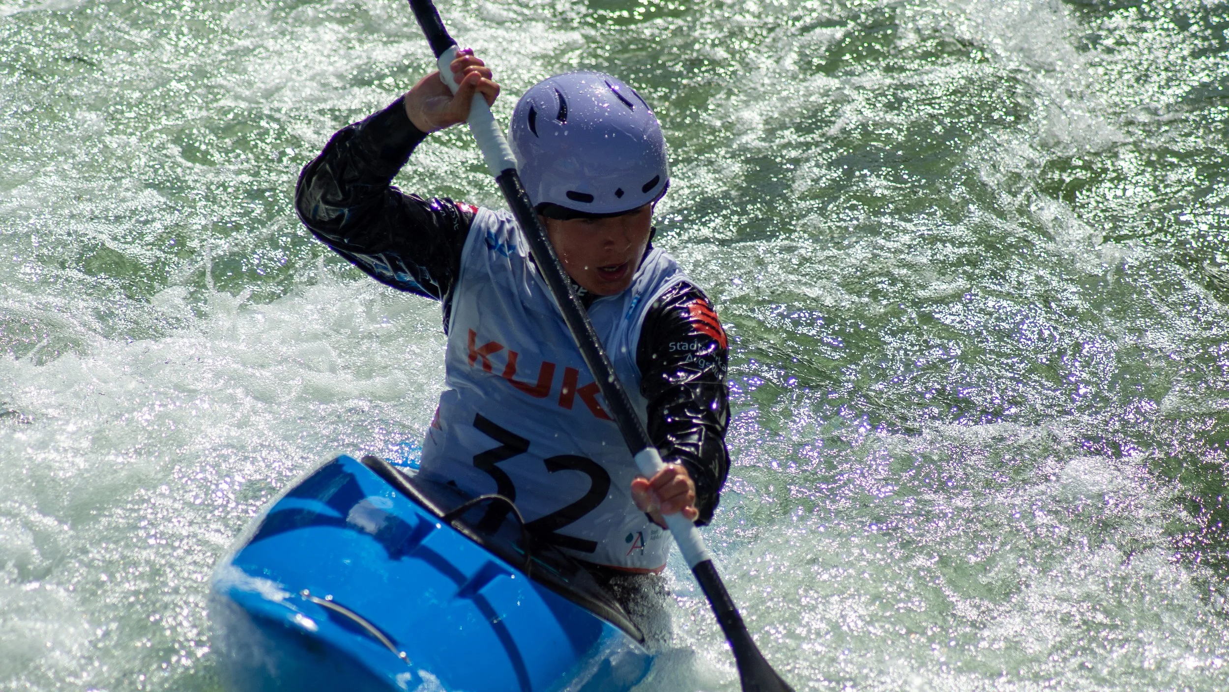 A person kayaking in rough water, wearing a black jacket, white helmet, and life vest with the number 32 on it.