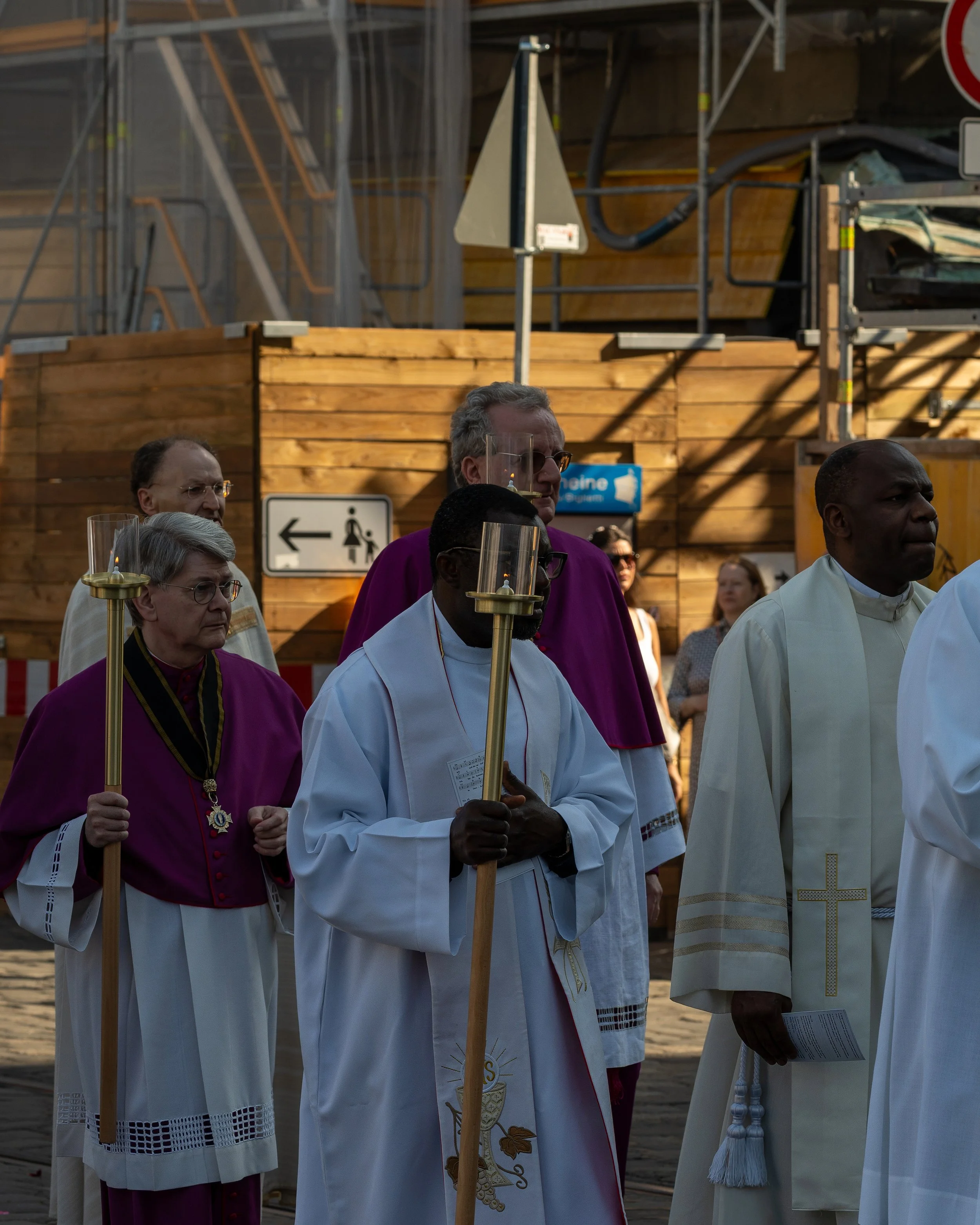 Clergy members dressed in purple and white robes participating in a religious procession outdoors, some holding lit candles, against a background of construction site elements and pedestrians
