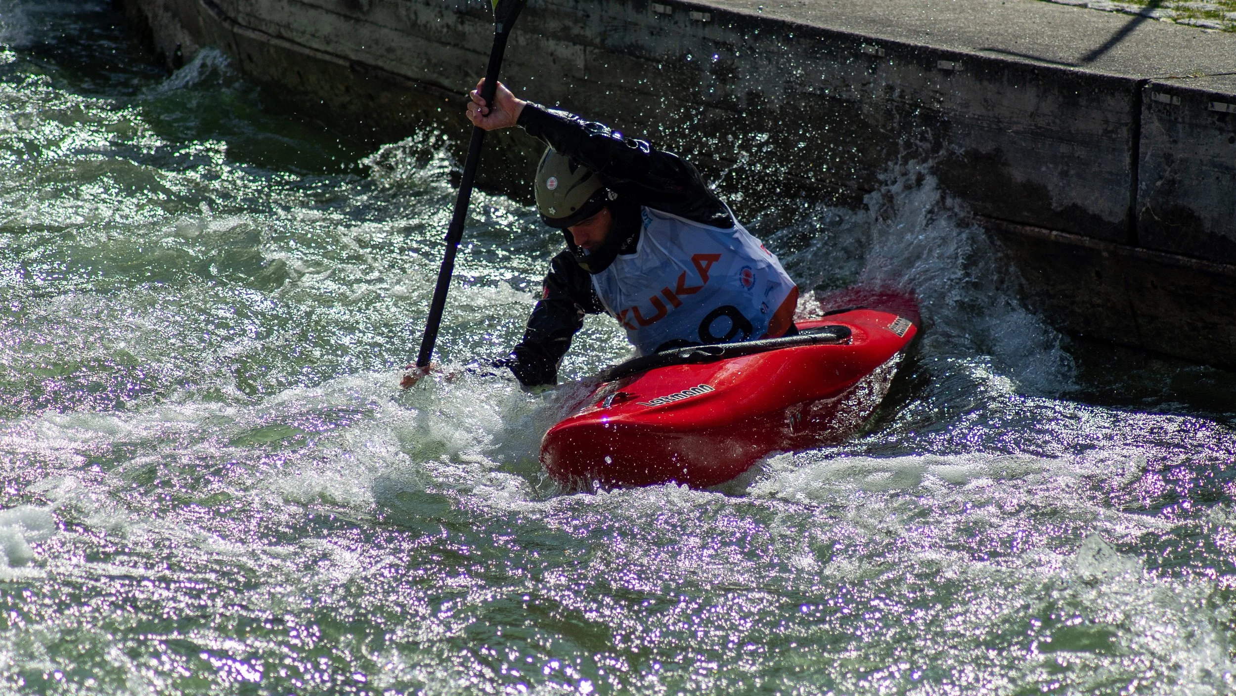 A kayaker wearing a helmet and a life jacket maneuvers through whitewater rapids near a concrete edge.