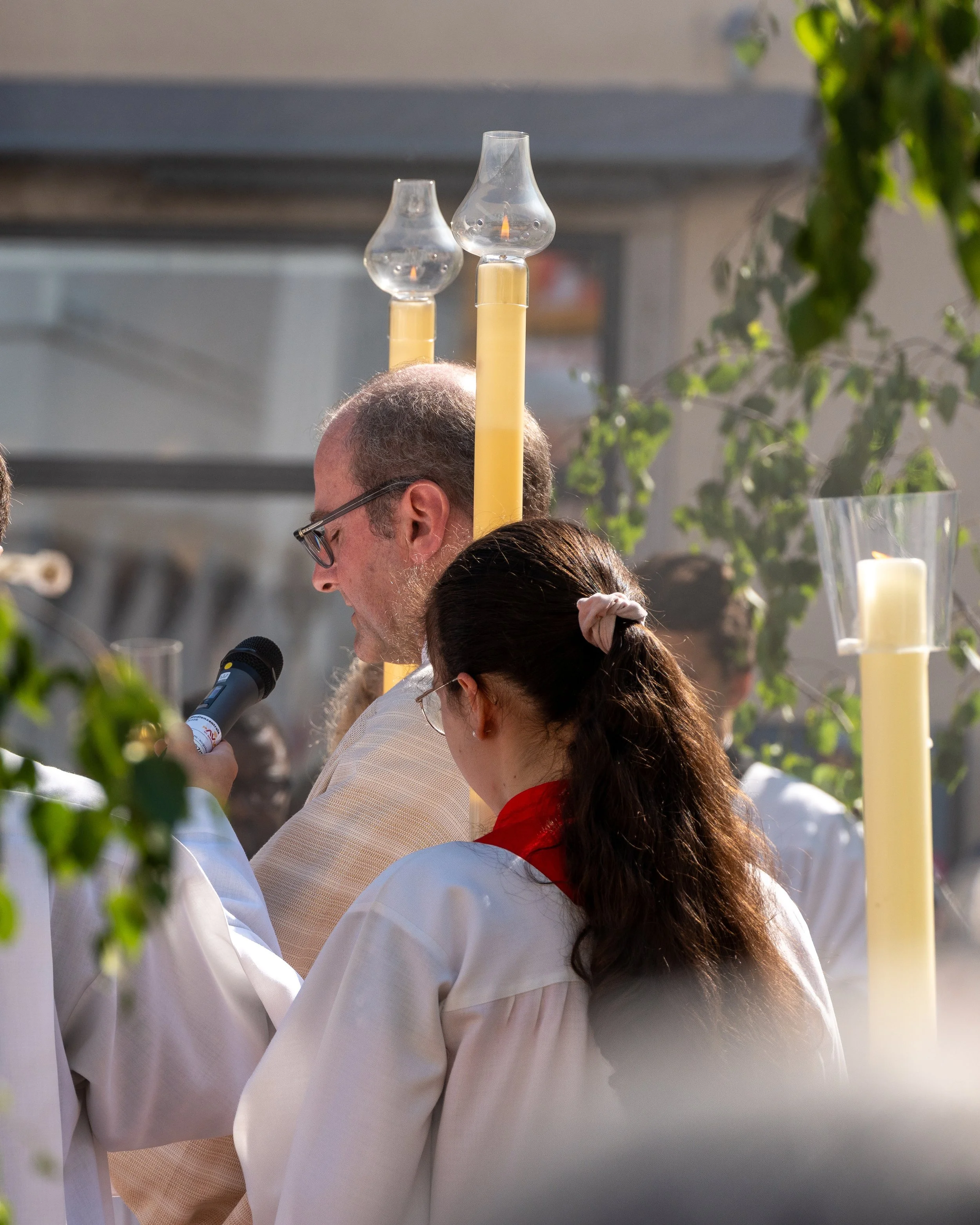 A man and a woman are participating in a religious ceremony outdoors, with the man holding a microphone and the woman looking down. There are tall candles and greenery surrounding them.