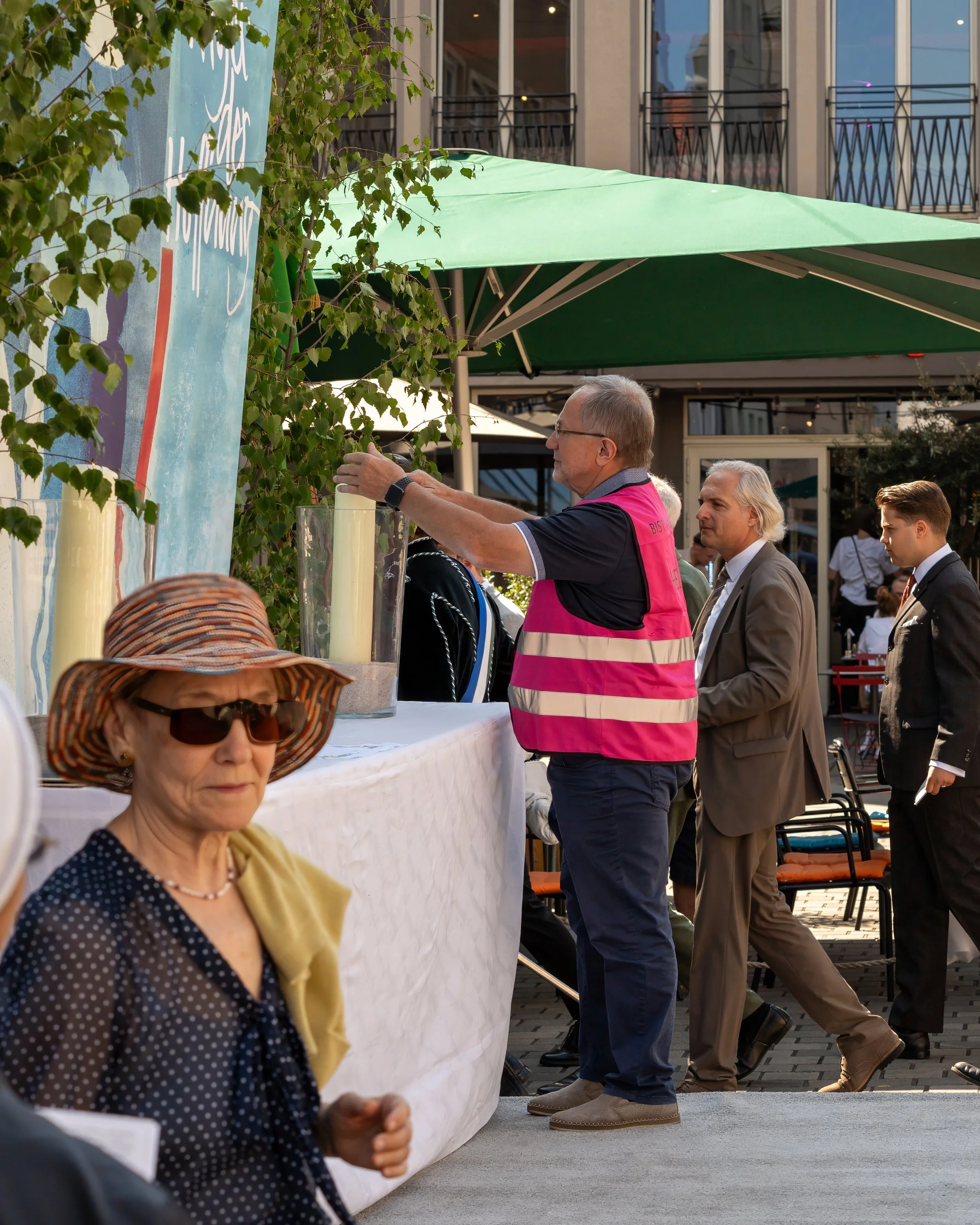 A man in a pink safety vest serves food or drinks at an outdoor event, with several well-dressed people standing around, including an elderly woman with sunglasses and a wide-brimmed hat in the foreground.