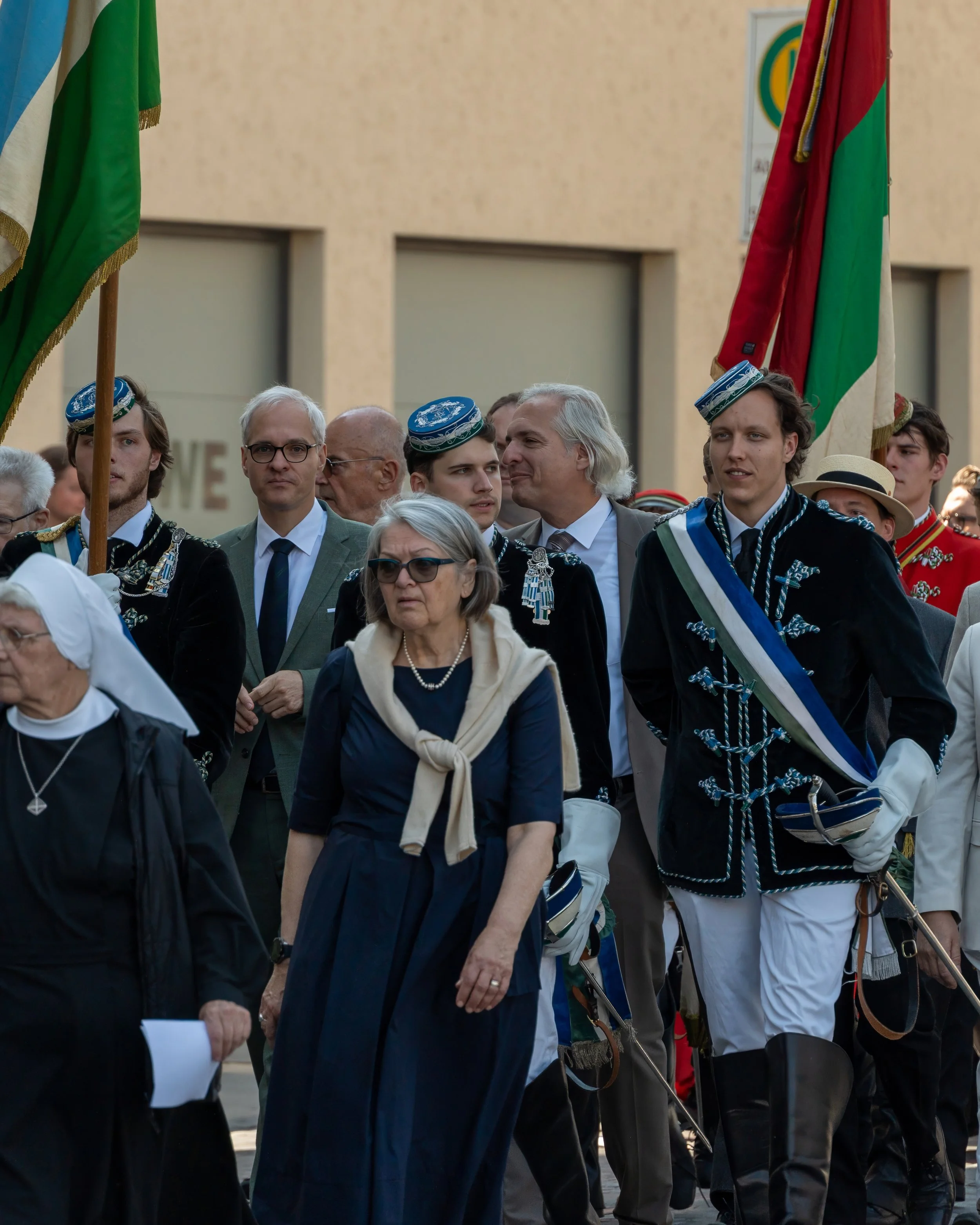 Group of people participating in a parade or procession, with some individuals dressed in traditional military or ceremonial uniforms, carrying flags, including green and red flags, with a beige building in the background.