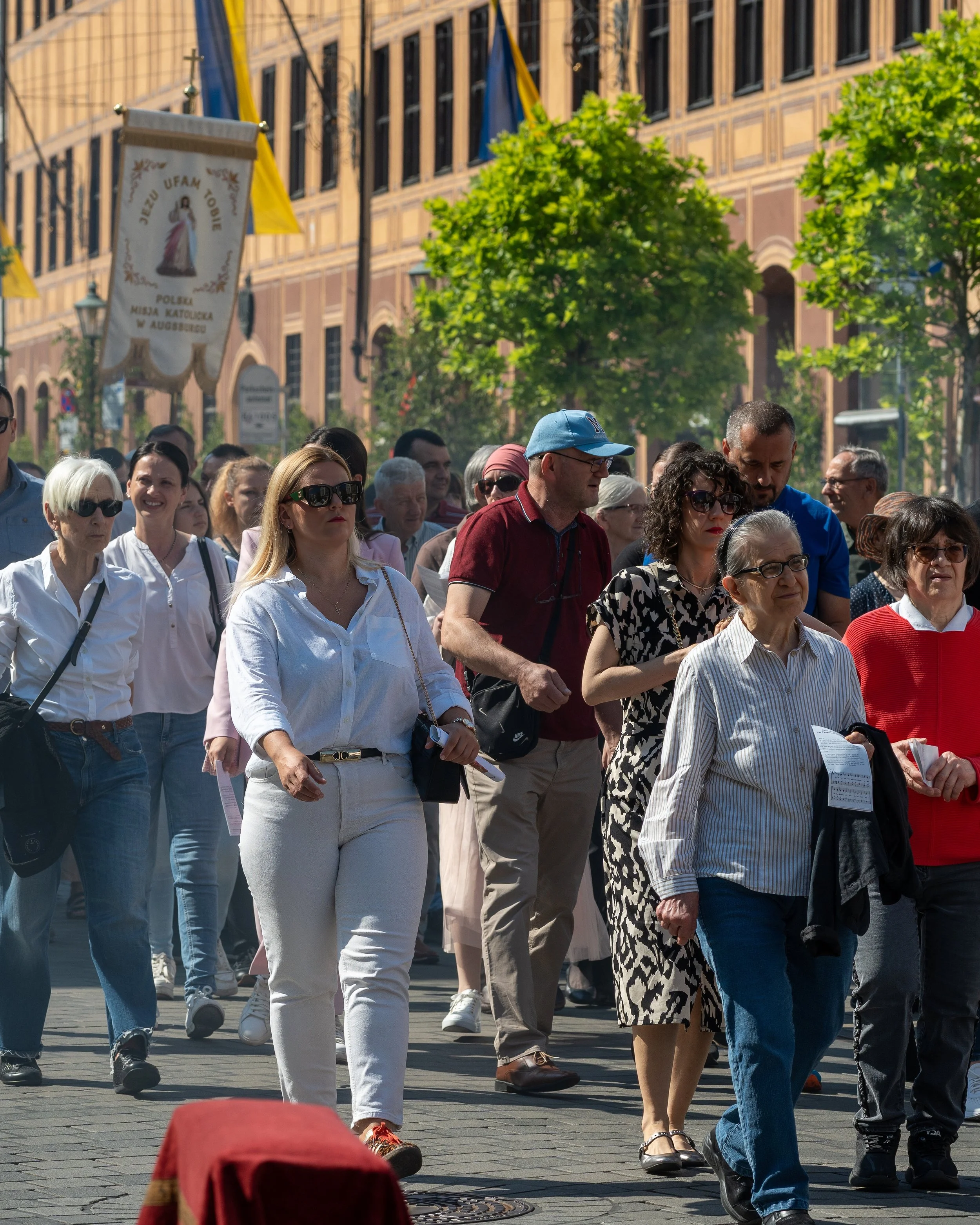 A diverse group of people walking together on a city street during daytime, with buildings, green trees, and yellow flags in the background.