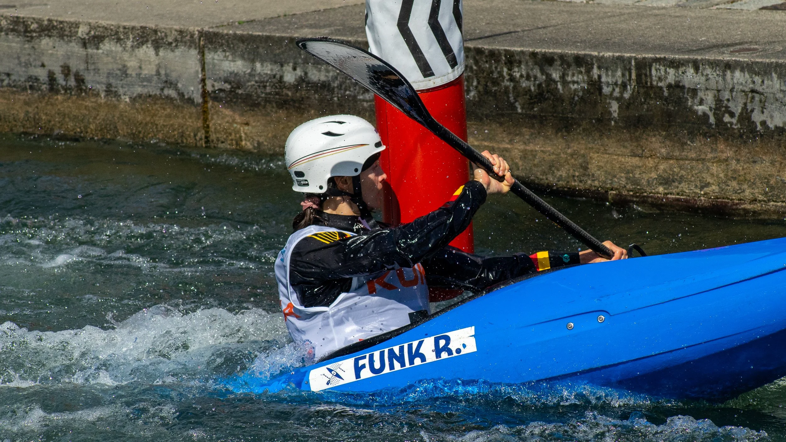 A person wearing a white helmet and black and white sportswear paddling a blue kayak on a river, with a red buoy and concrete bank in the background.