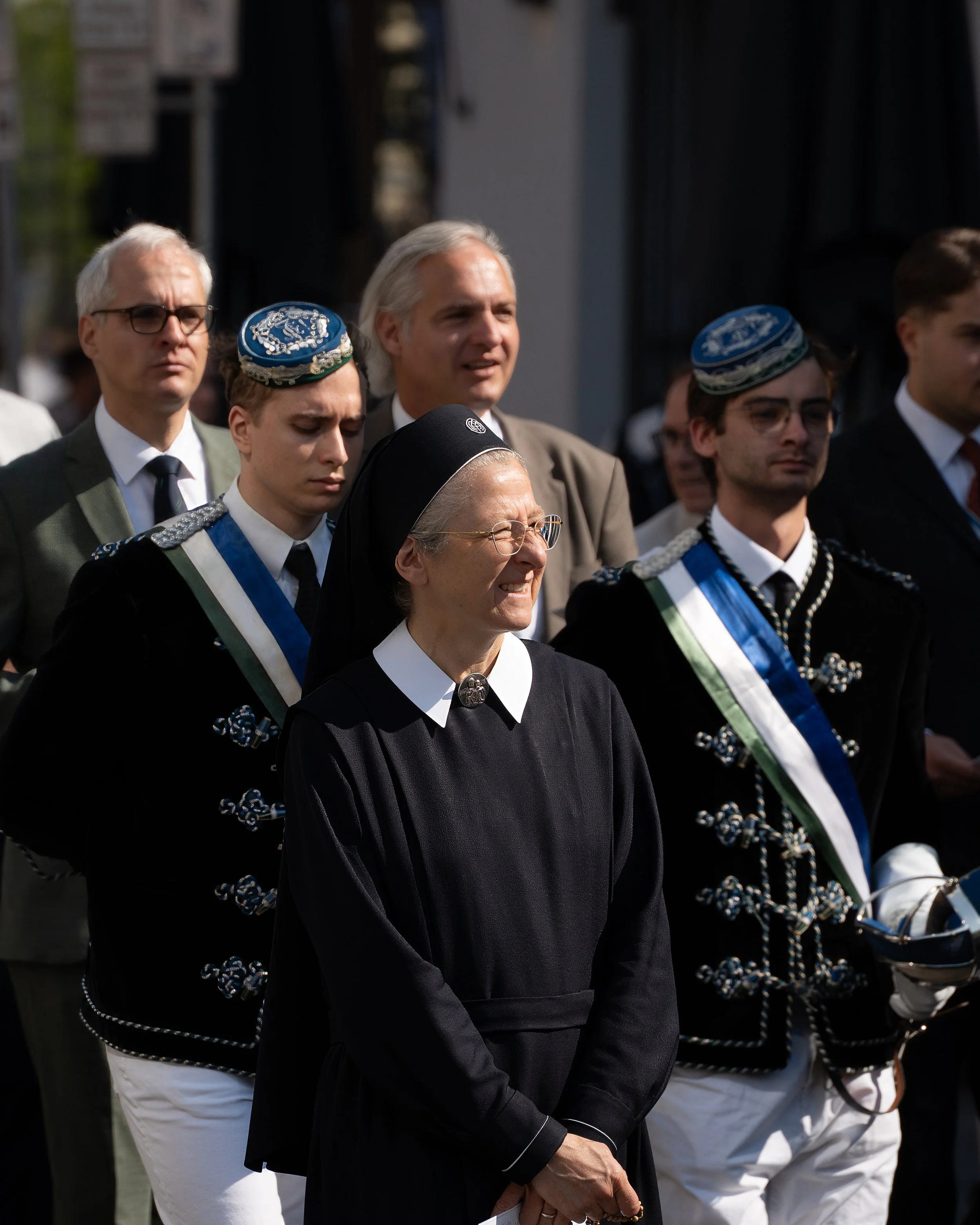 A group of people, including a nun and men in traditional Nordic costumes, attending an outdoor event, with some looking solemn and others smiling.
