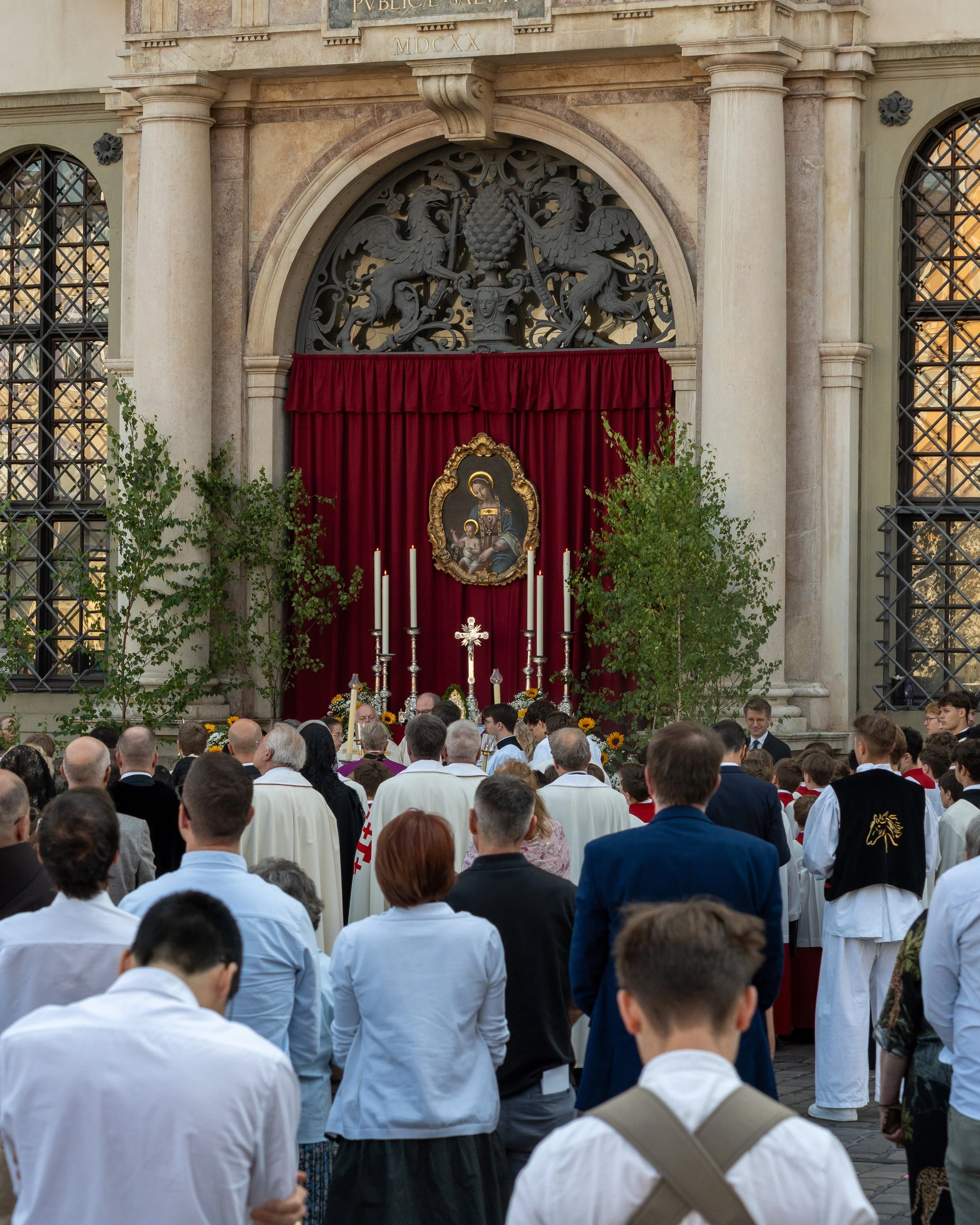 A religious gathering outside a church, with people attending a ceremony in front of an altar featuring a painting of the Madonna and Child, candles, and a red curtain, framed by two small trees and architectural decorations.