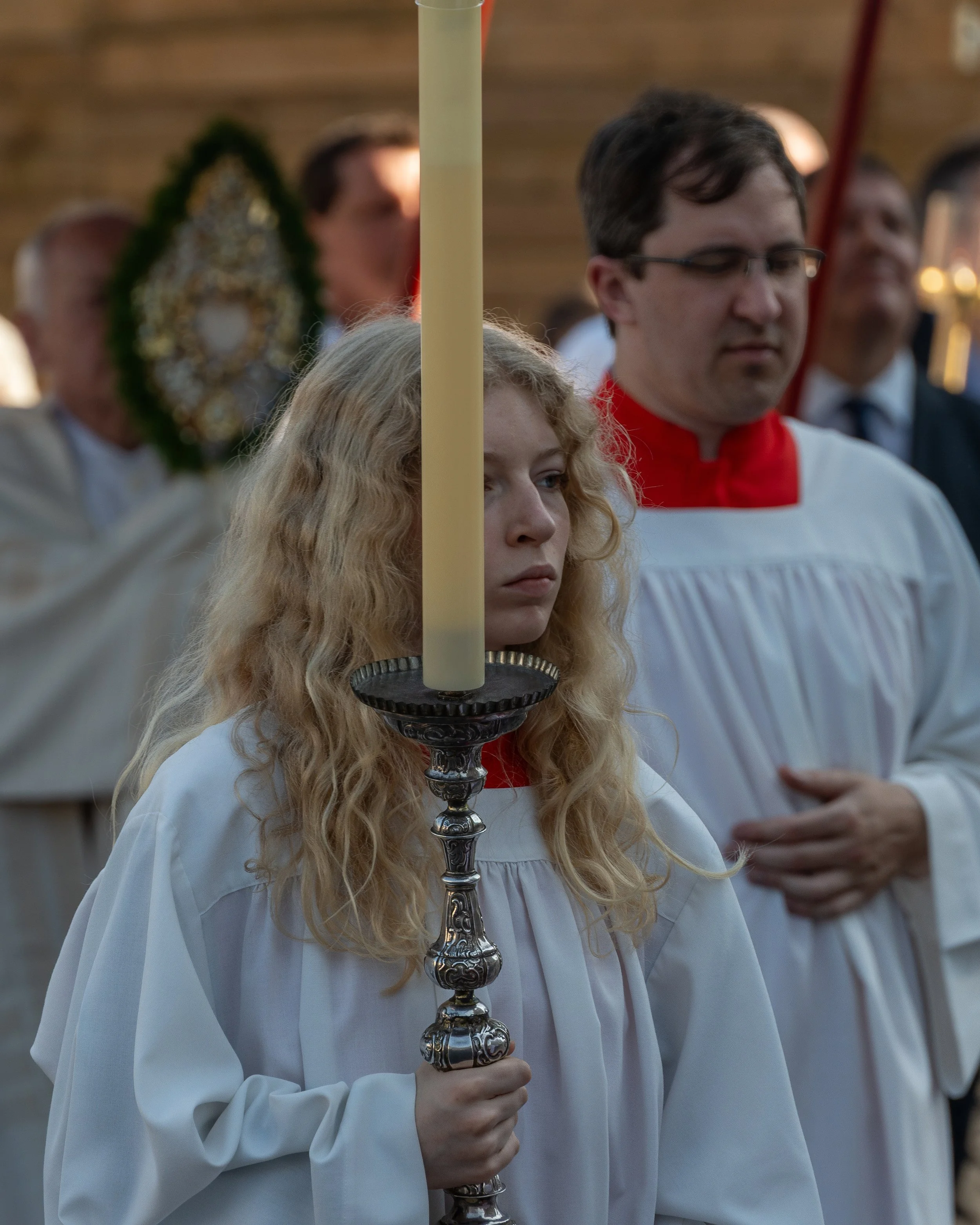 Young girl with long curly blonde hair holding a lit candle during a religious procession or ceremony.