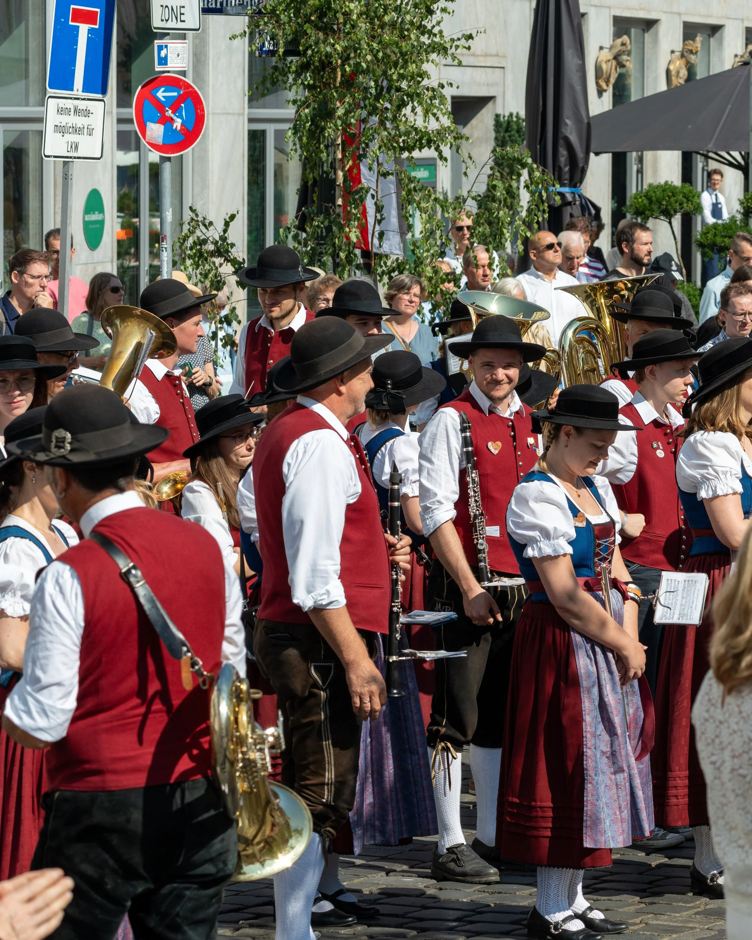 Group of people dressed in traditional Bavarian clothing participating in a parade, some holding musical instruments, on a cobblestone street with onlookers and signs in the background.