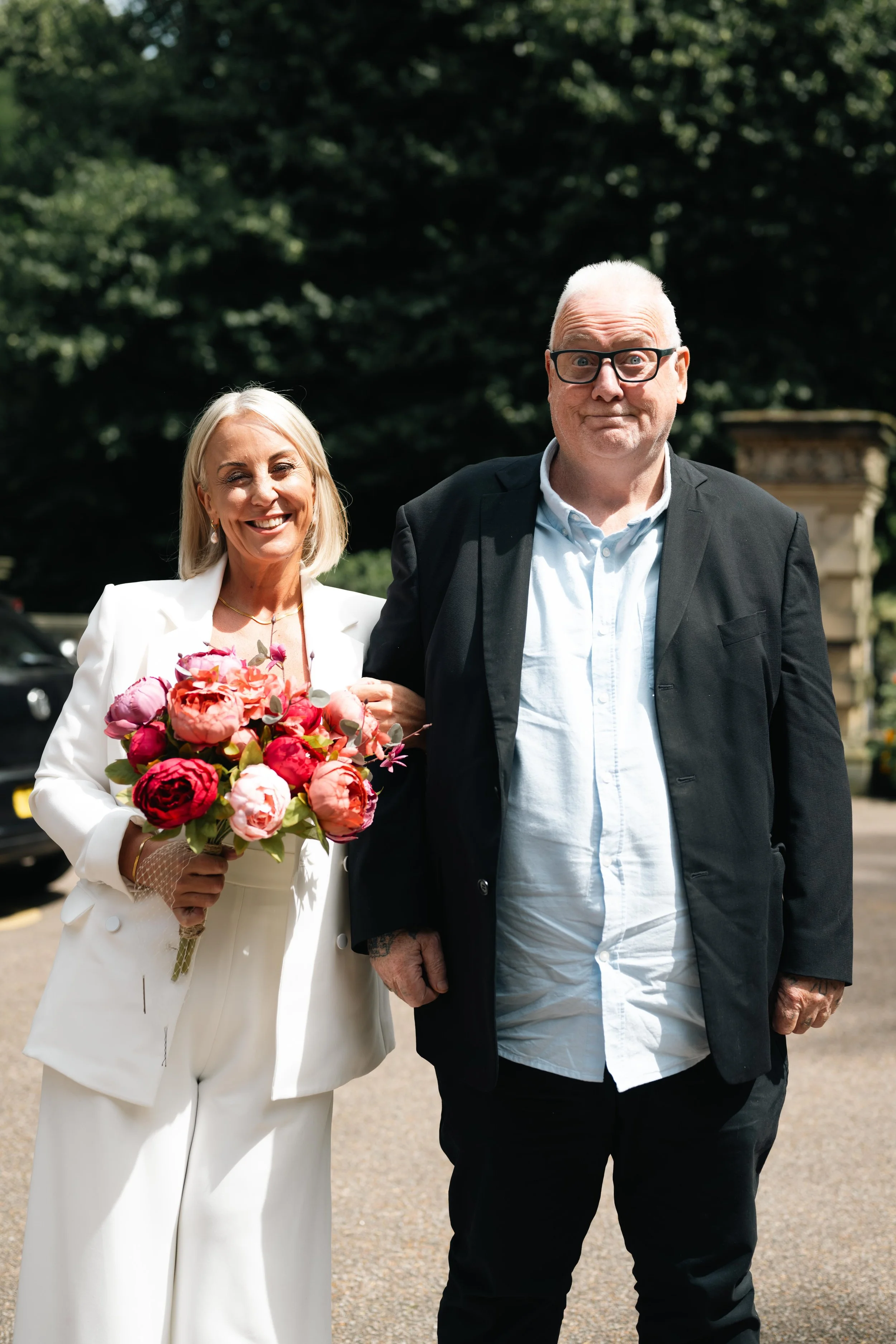 A woman in a white suit holding a bouquet of pink and red flowers, standing next to a man in a black jacket and white shirt outdoors on a sunny day, with trees in the background.