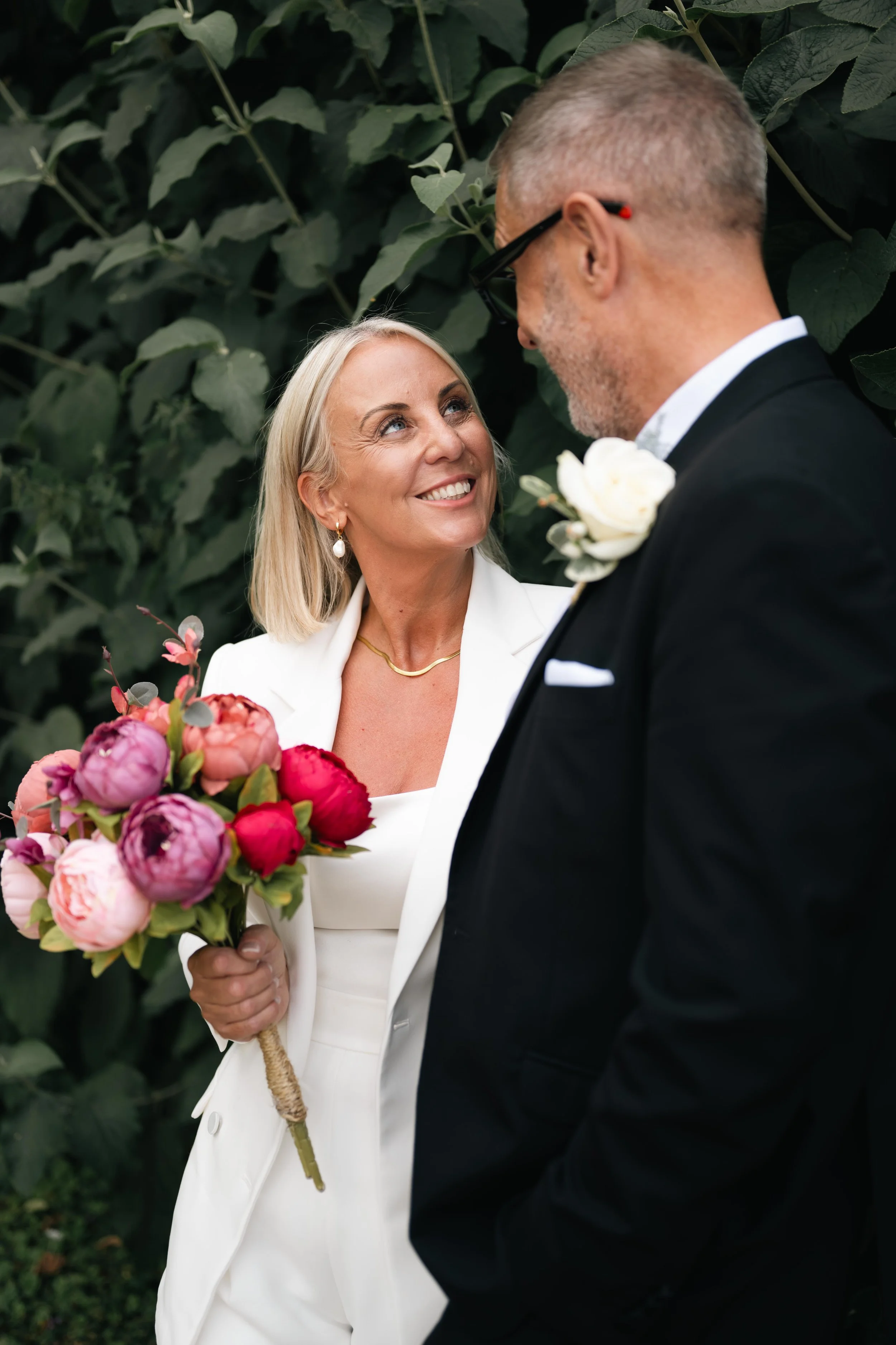 A woman in a white outfit holding a colorful bouquet, smiling up at a man in a black suit with a white boutonniere, in front of green foliage, at a wedding or special celebration.