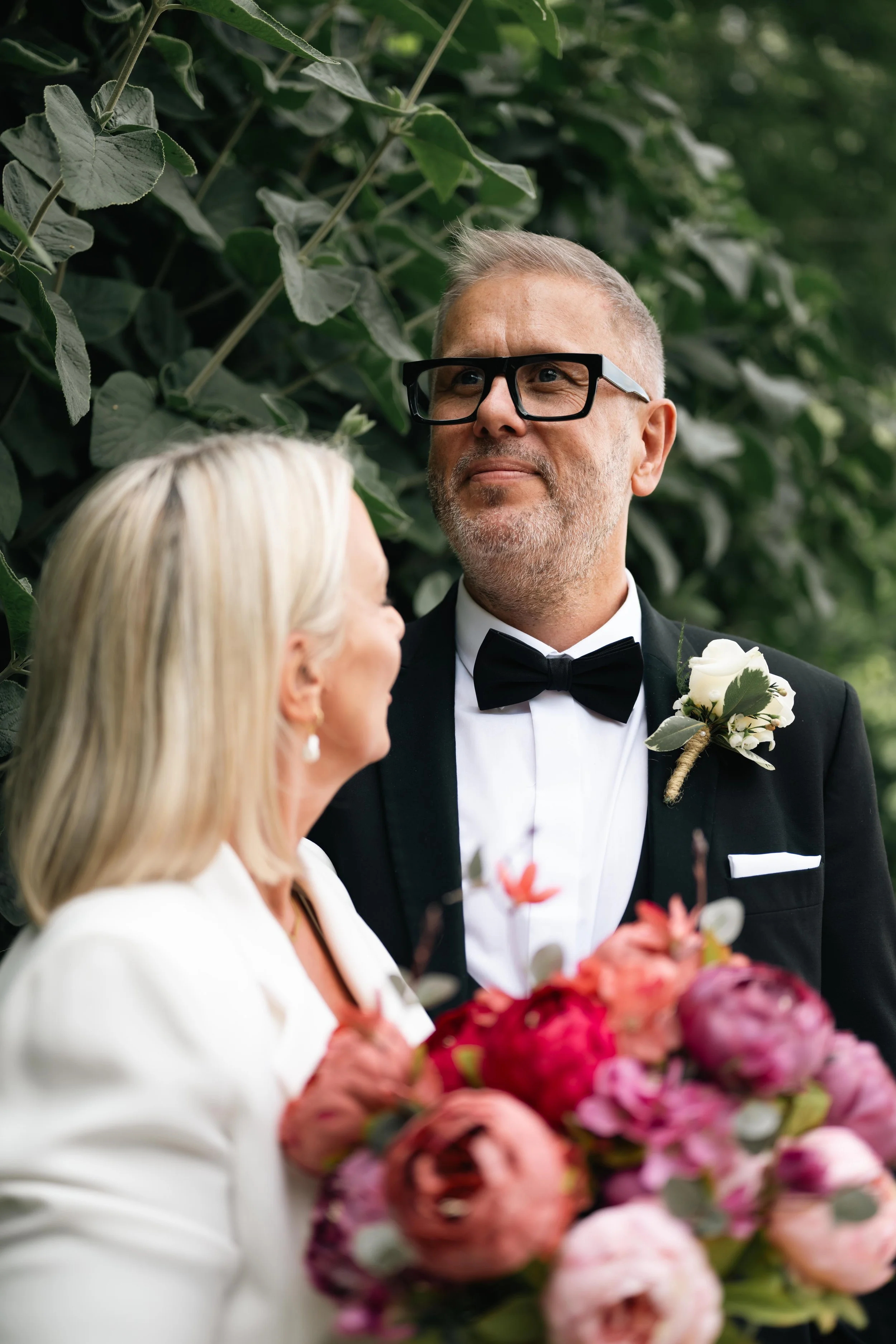 A bride holding a bouquet of pink and purple flowers standing next to a groom dressed in a black tuxedo with a white shirt, black bow tie, and a white boutonniere, outdoors with green foliage in the background.