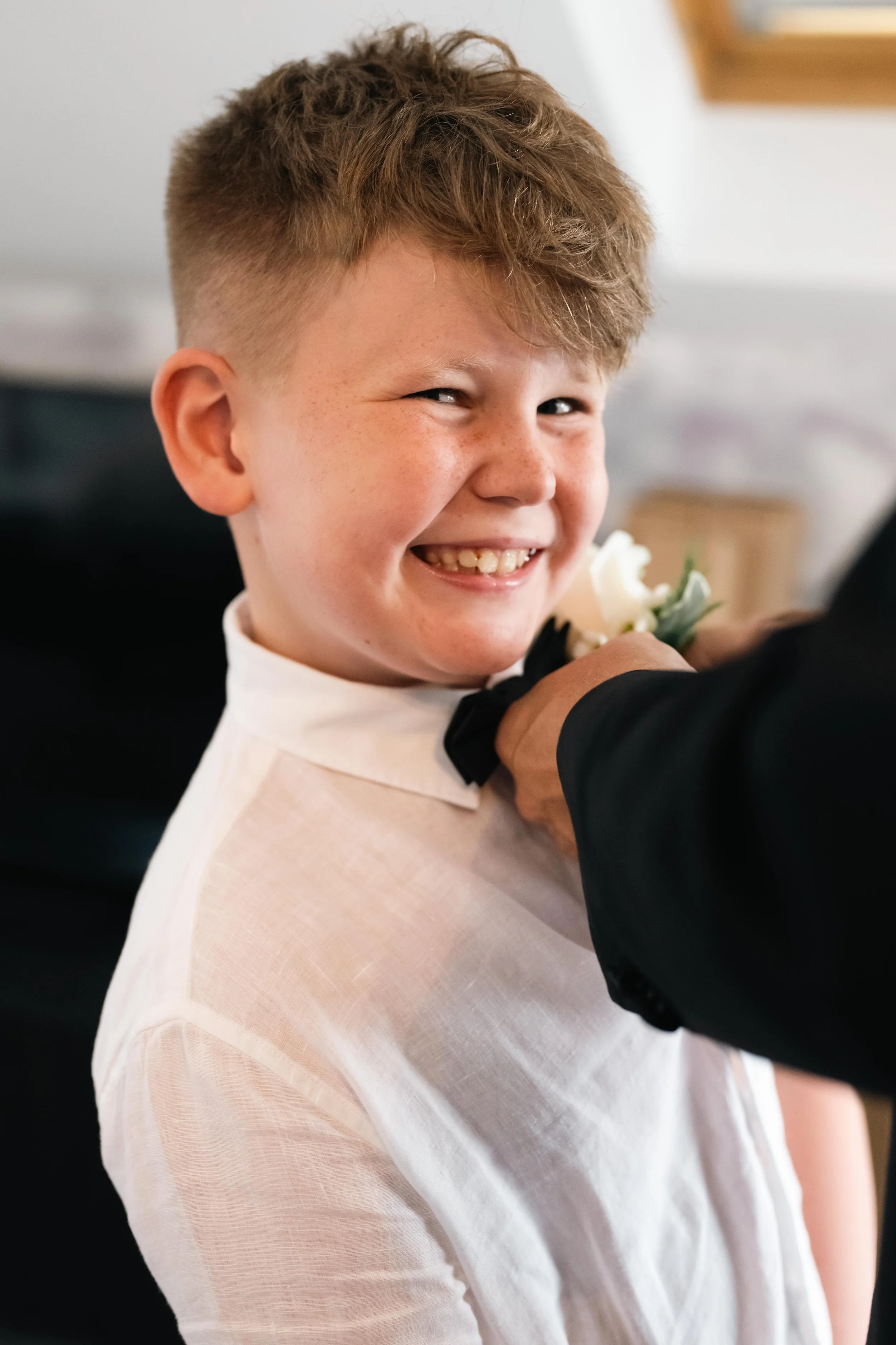 A young boy with curly red hair and freckles wearing a white shirt and a black bow tie, smiling as he adjusts his bow tie in front of a person in a black suit.