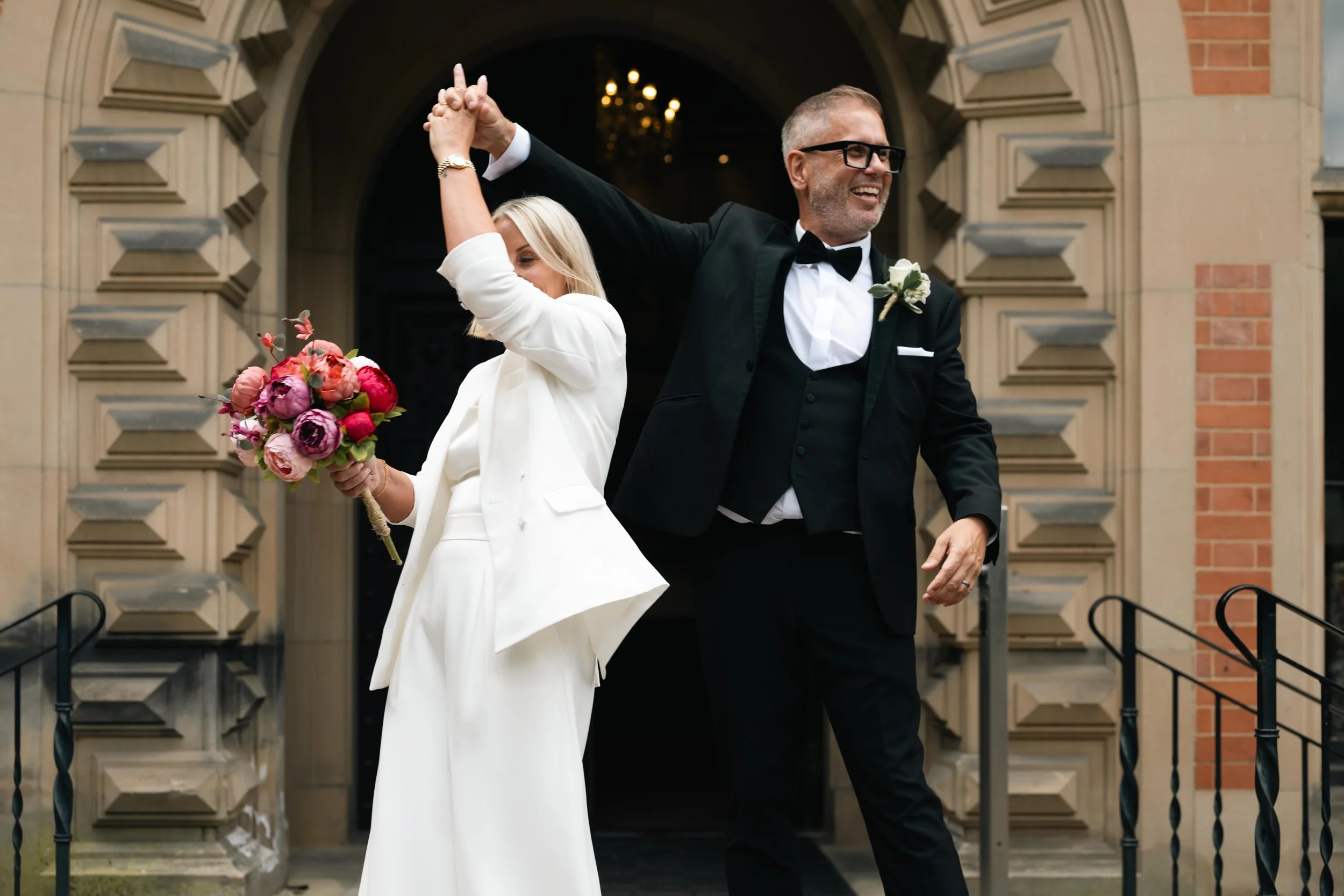 Bride and groom share a dance outside a church, holding hands in a twirl, with the bride holding a bouquet of pink and purple flowers.