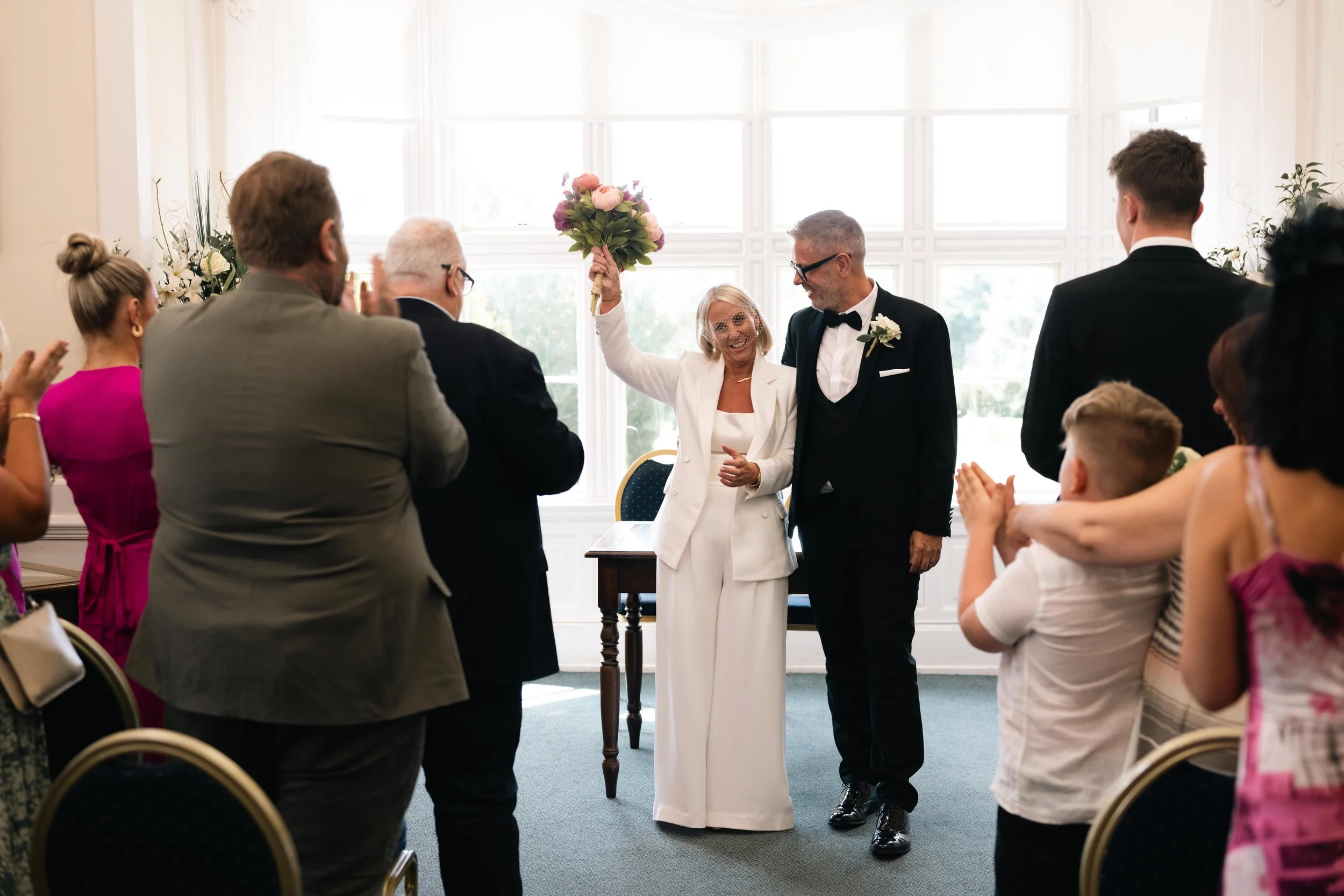 A wedding ceremony scene with a bride and groom standing in front of a large window, the bride holding a bouquet of pink flowers and all guests clapping, celebrating.