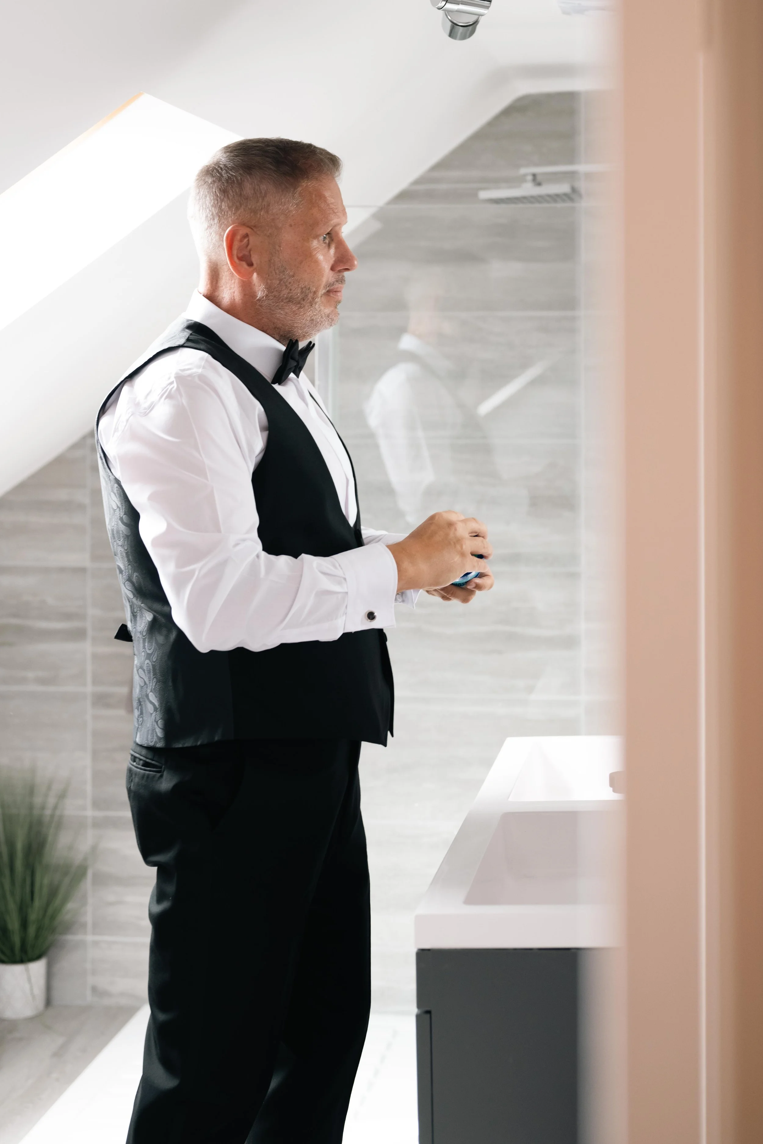 Man in tuxedo with bow tie standing in front of mirror in modern bathroom.