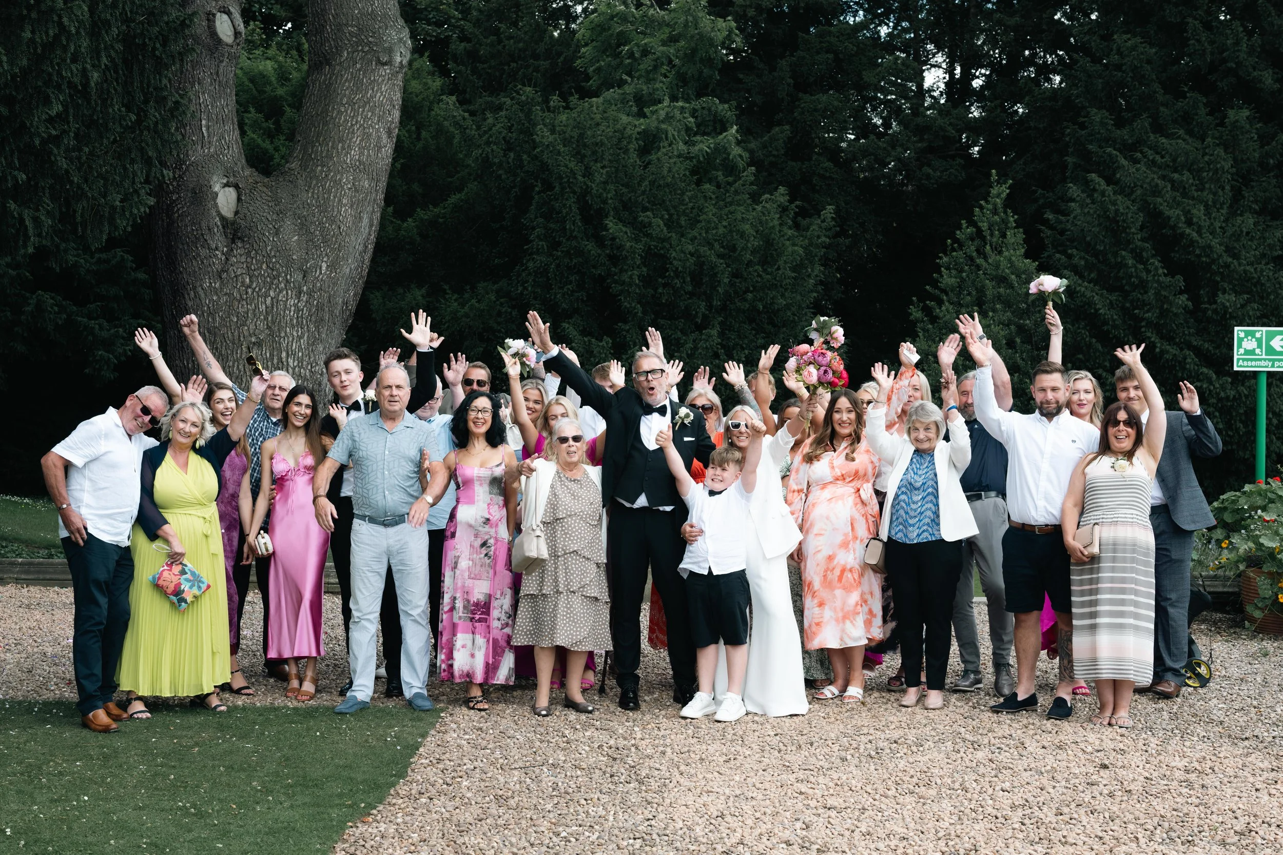 Group of people celebrating at a wedding outdoors, smiling with arms raised, with a large tree and green foliage in the background.