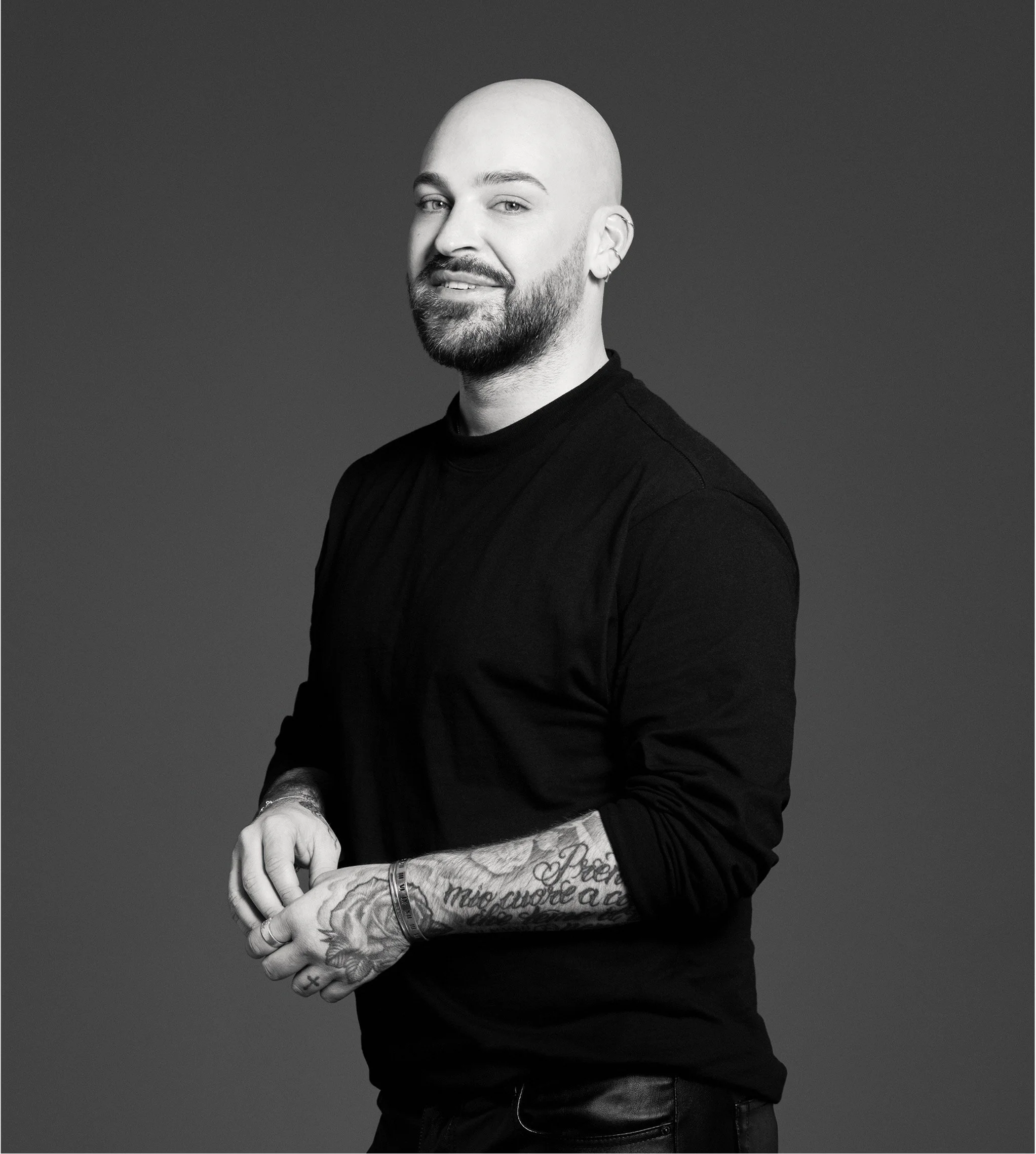 Schwarze und weiße Porträtaufnahme eines Mannes mit Glatze, Bart und Tätowierungen am Arm, trägt schwarzes Langarmshirt, lächelt leicht, vor grauem Hintergrund.