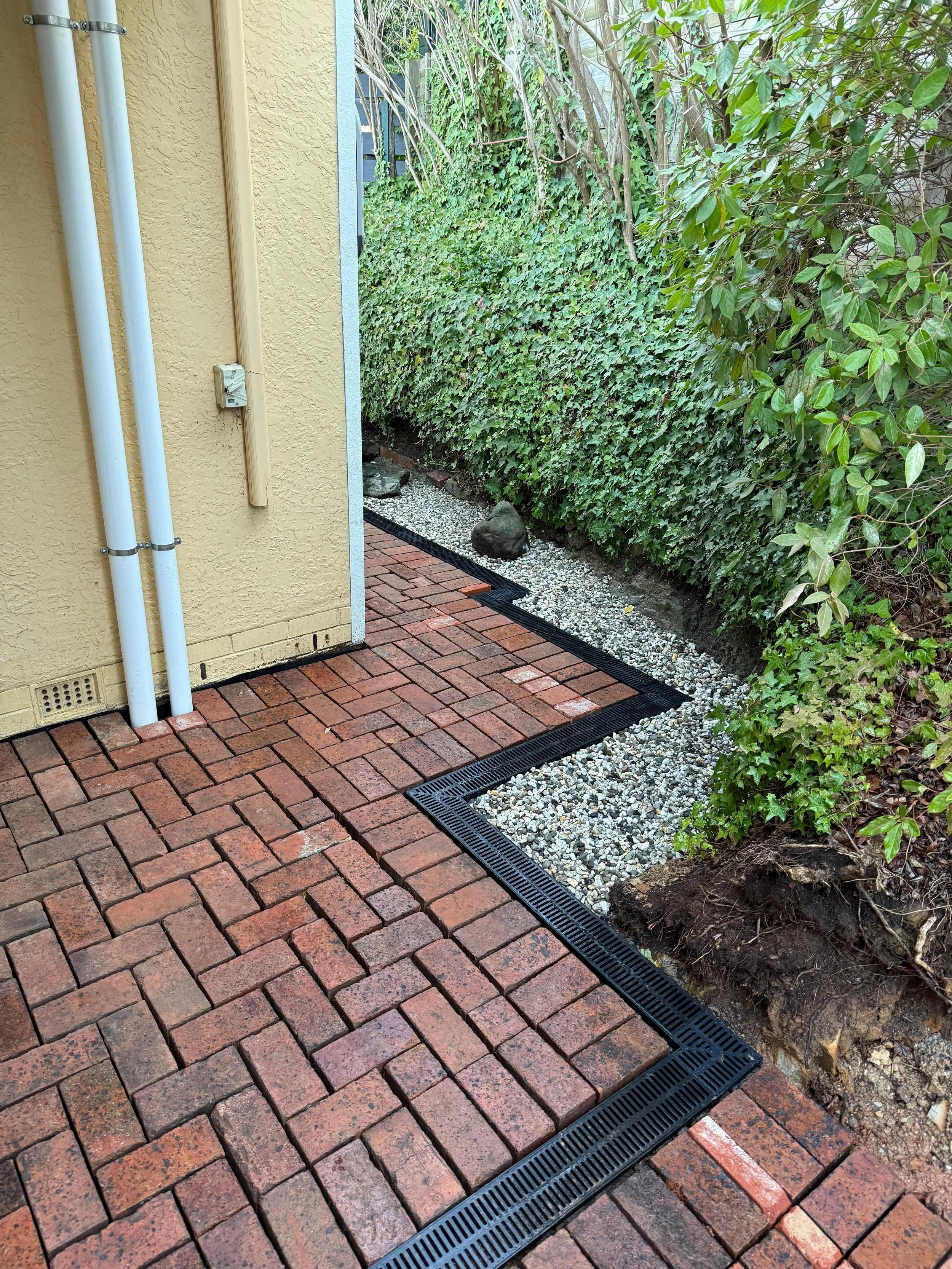 Brick patio with metal drainage grate along the edge, next to a garden with rocks and lush green plants.