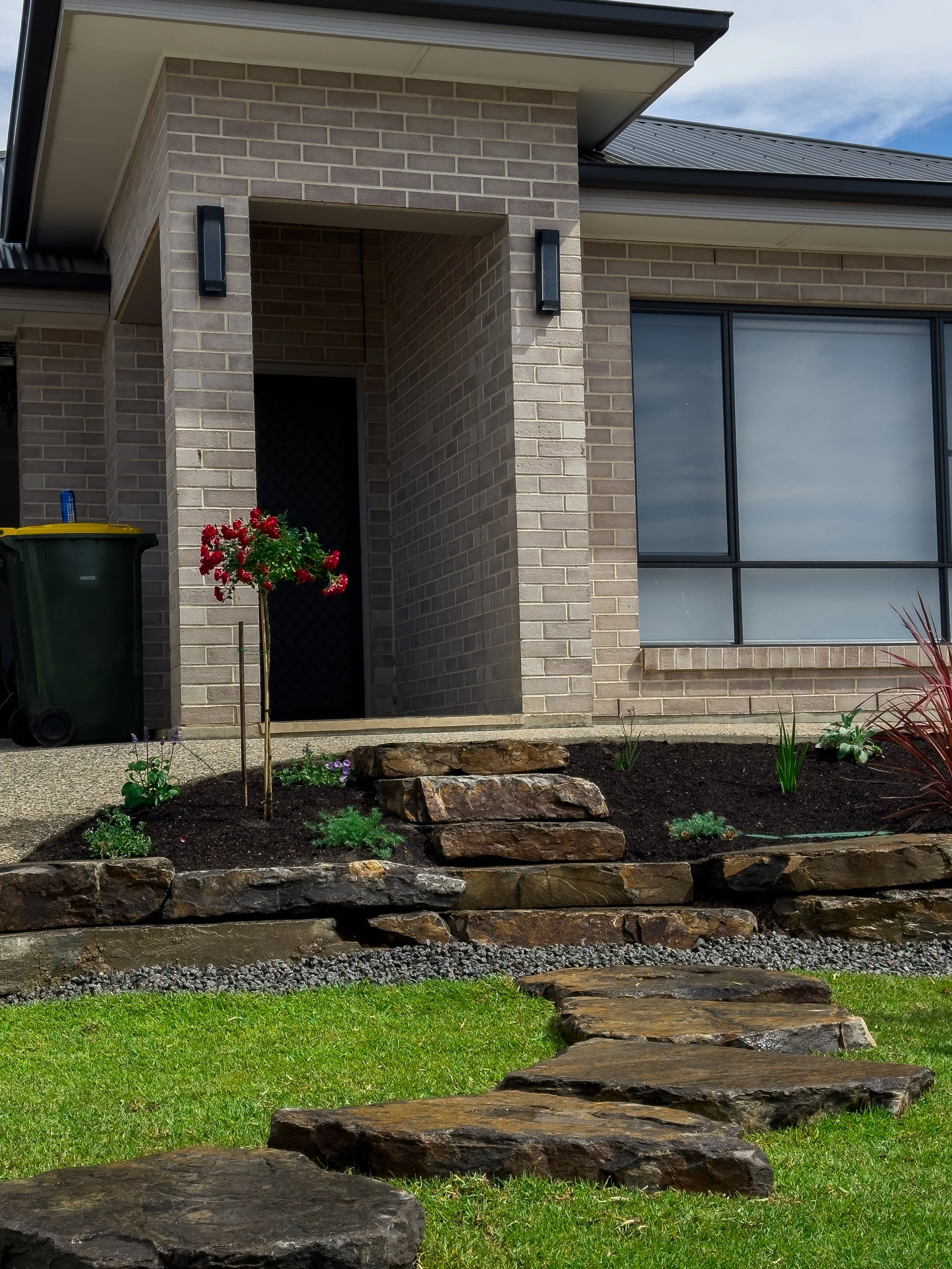 Front lawn with stone pathway, flower bed with plants, brick house entrance, window, and trash bin.