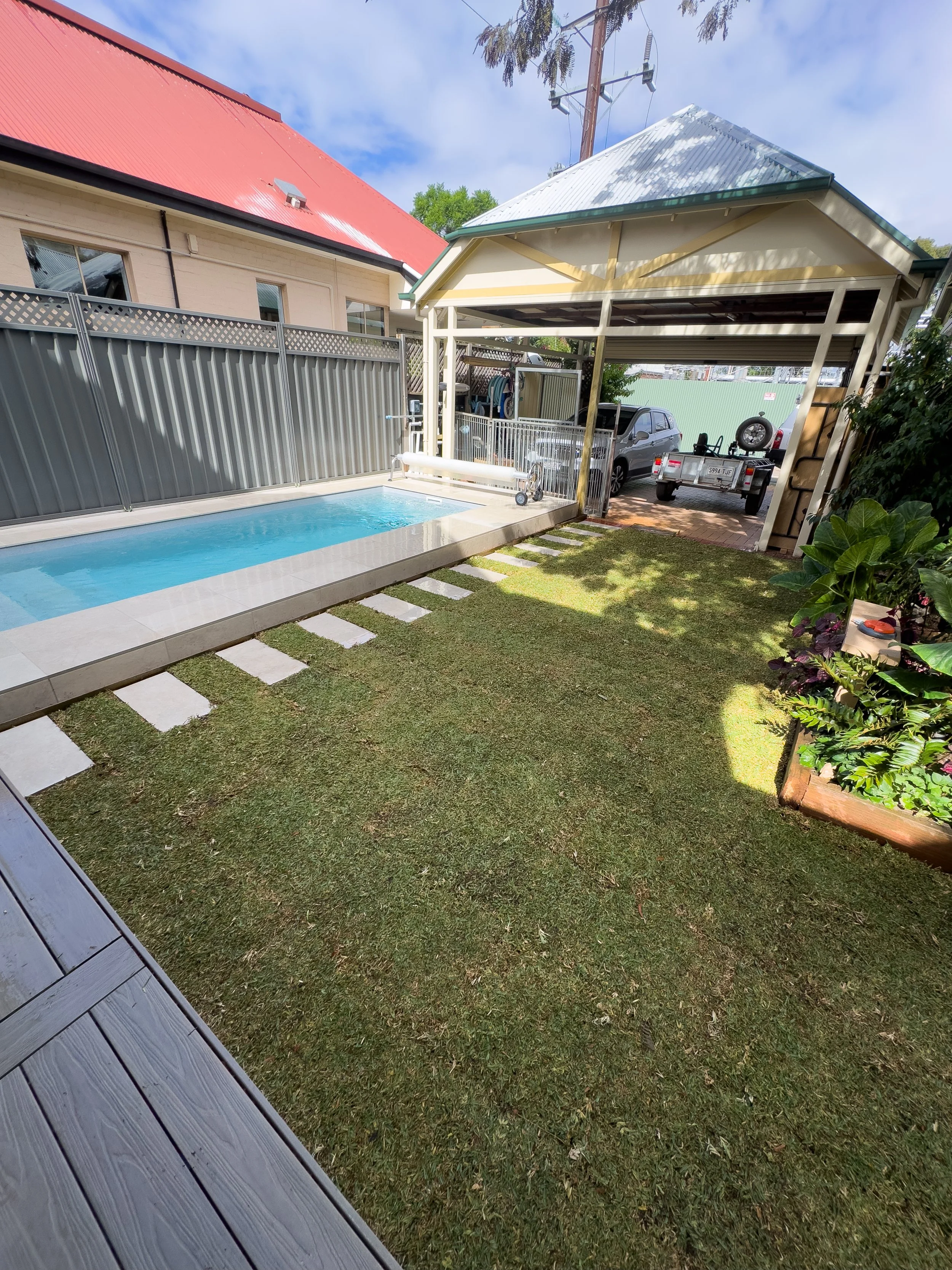 Backyard with a small swimming pool, a grassy lawn, a shed with a car and trailer parked inside, and a wooden deck part visible in the corner.