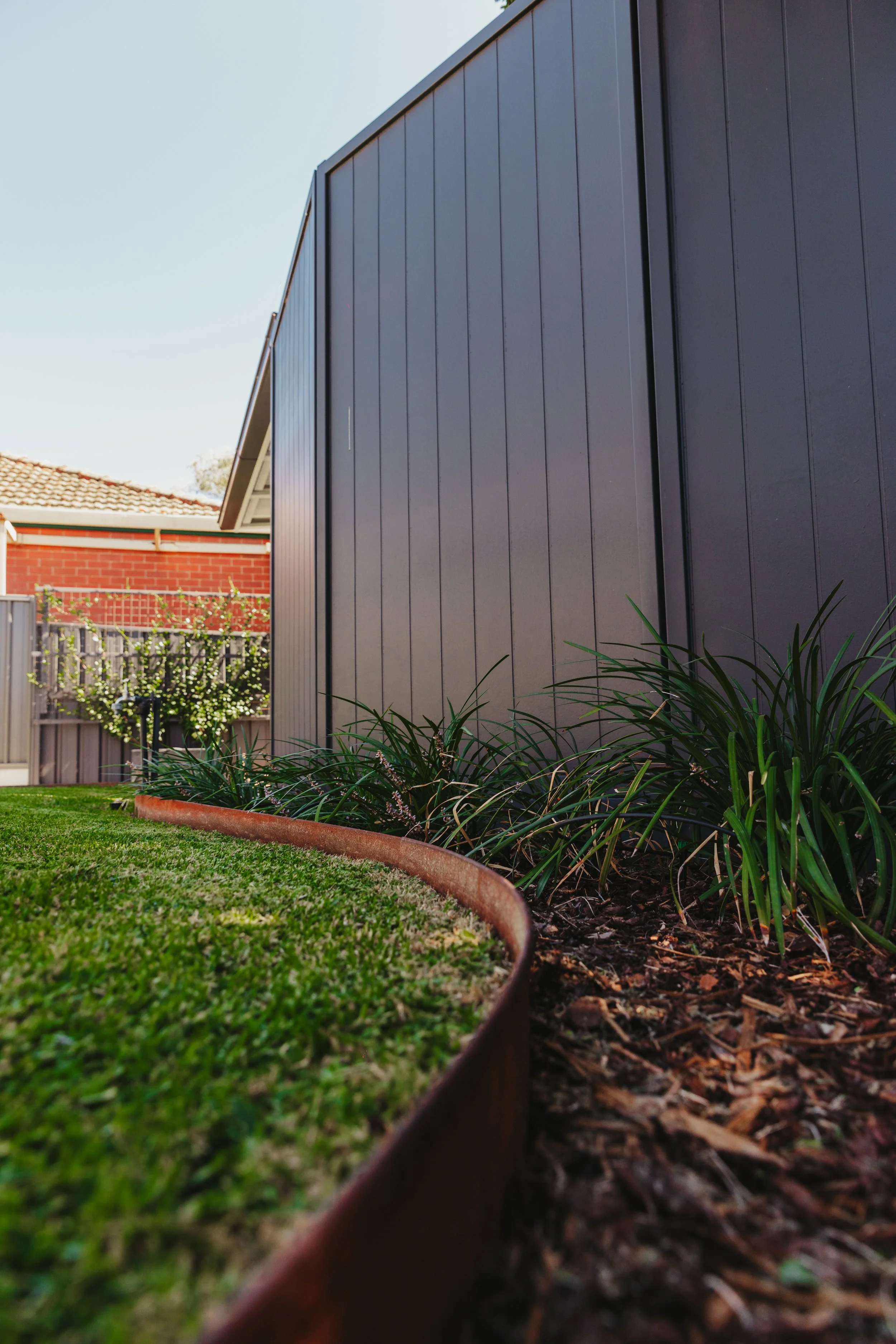 Side yard of a house with a gray metal fence, green grass, and plants along the base of the fence, with a brick house and bushes in the background.