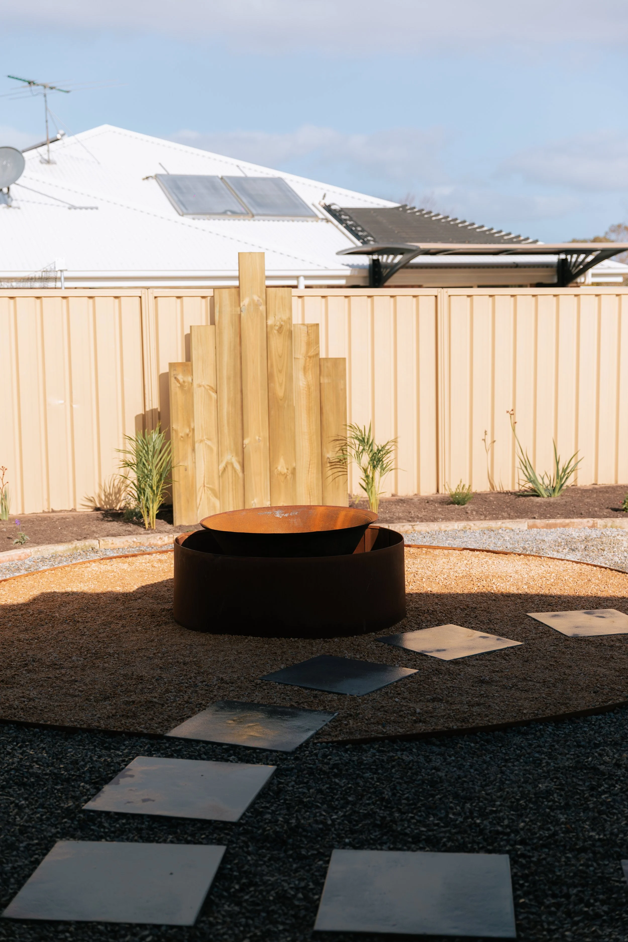 Backyard with a fire pit, stepping stones, and a beige fence, with a white house with solar panels and a rooftop outdoor structure in the background.