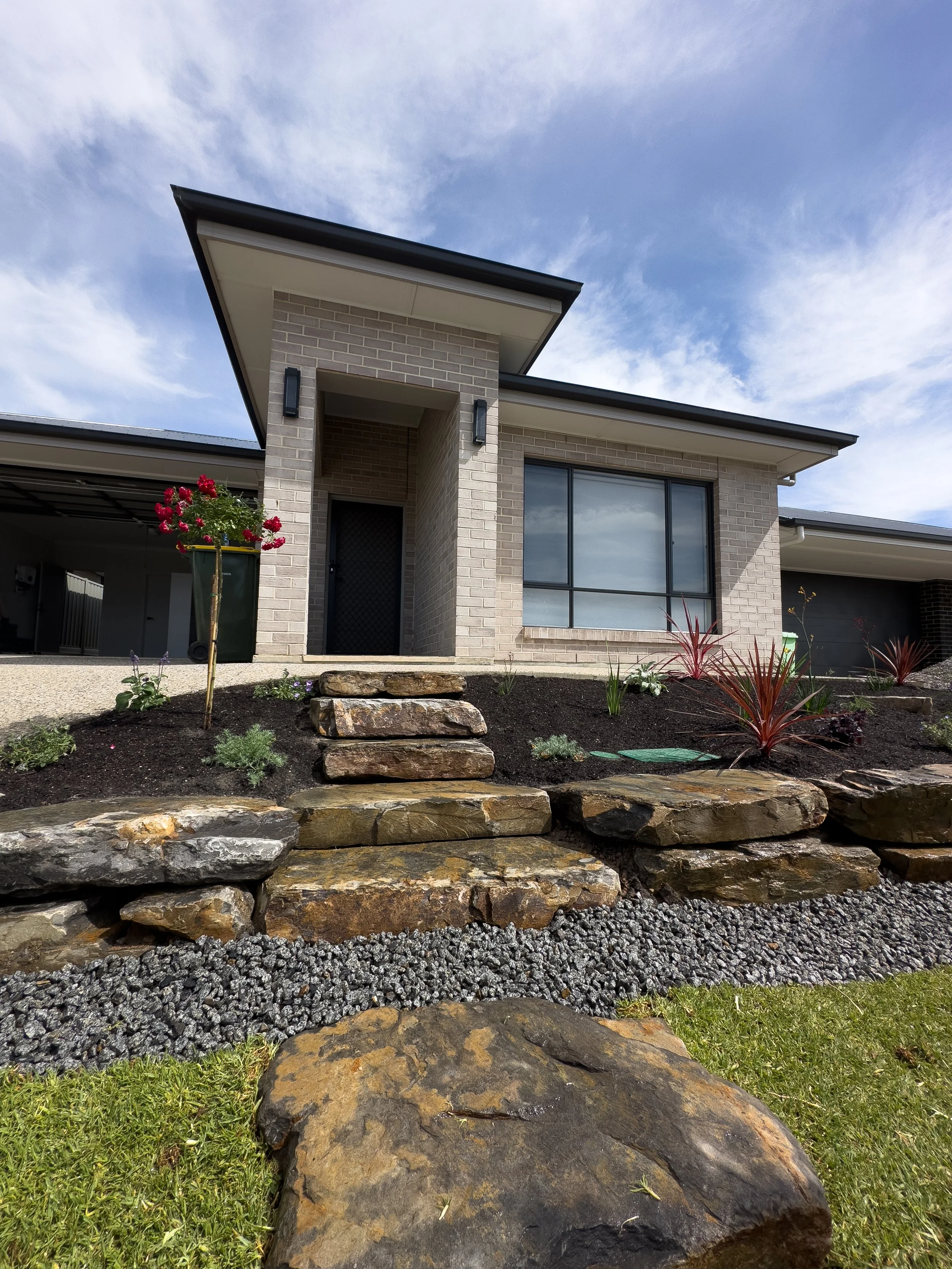 Front view of a modern house with a brick facade, large window, and landscaped yard with rocks, plants, and a small walkway.