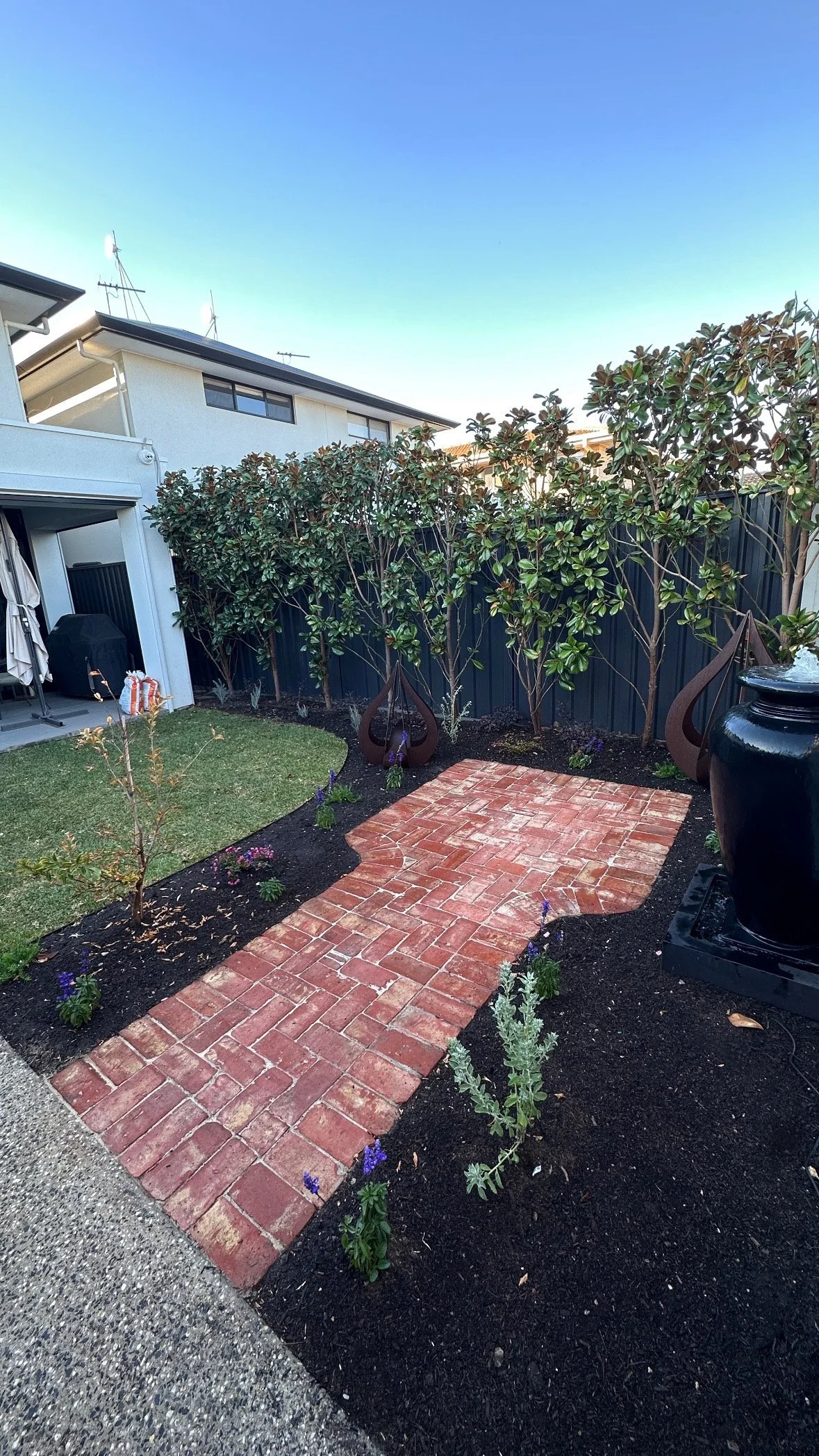 A small backyard garden with a brick pathway, young plants and shrubs, and large decorative metal sculptures against a dark fence.