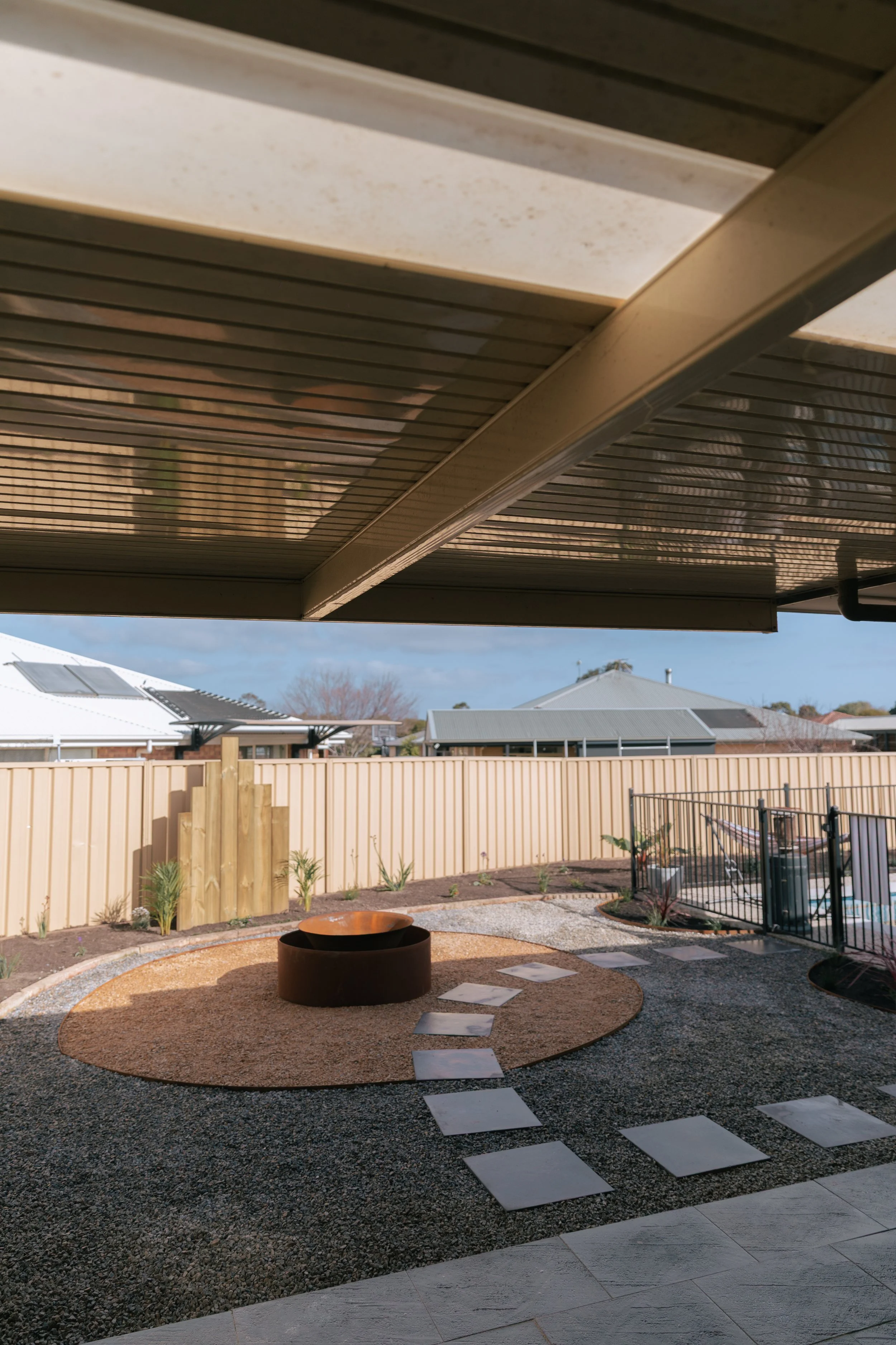Residential backyard patio with a fire pit, stepping stones, small plants, and a beige fence. Partially shaded by a roof structure.