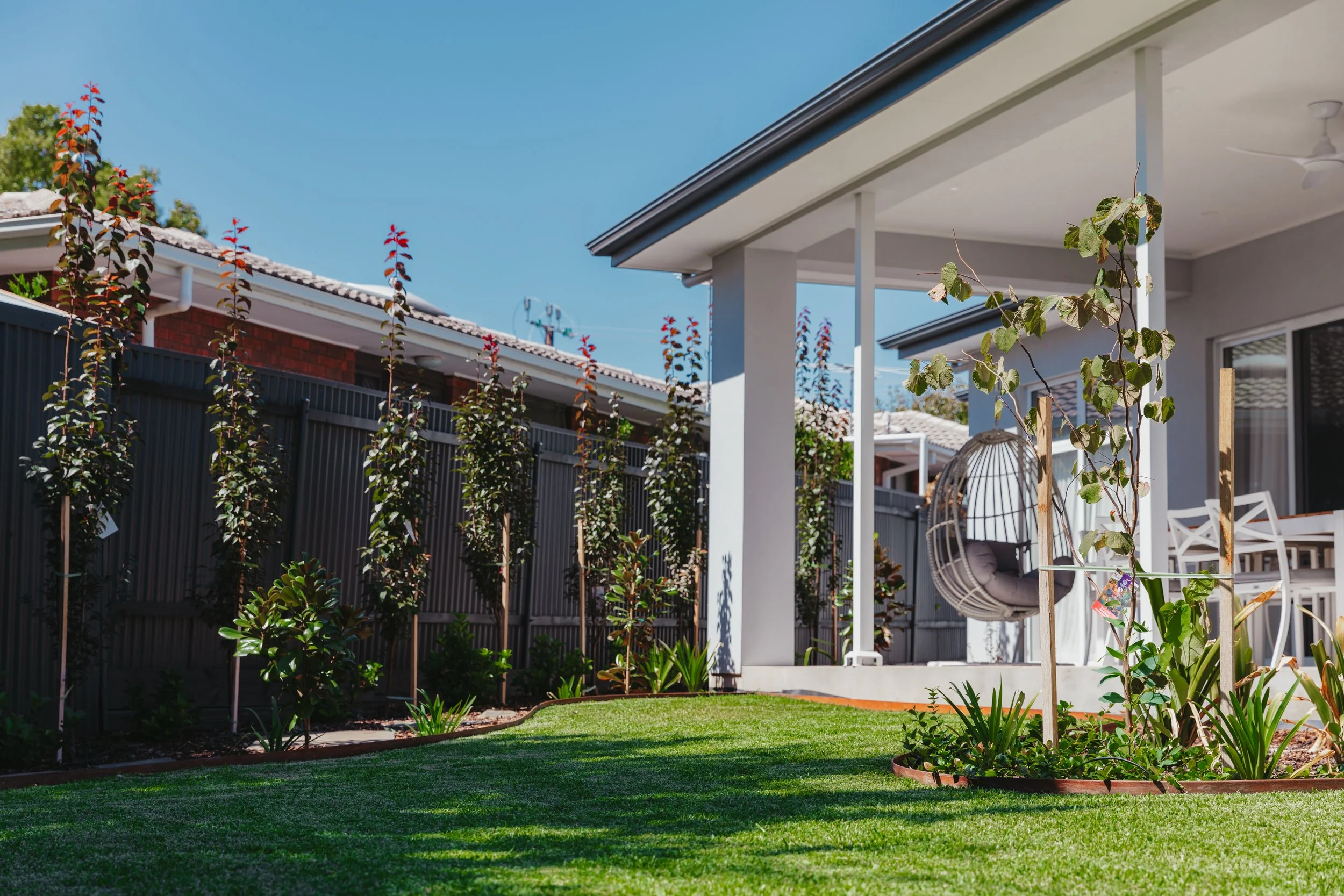 A backyard with a green lawn, small trees, and plants surrounded by a dark metal fence. There is a covered patio area with outdoor furniture, including a hanging egg chair, and a white dining table with chairs. The sky is clear and blue.