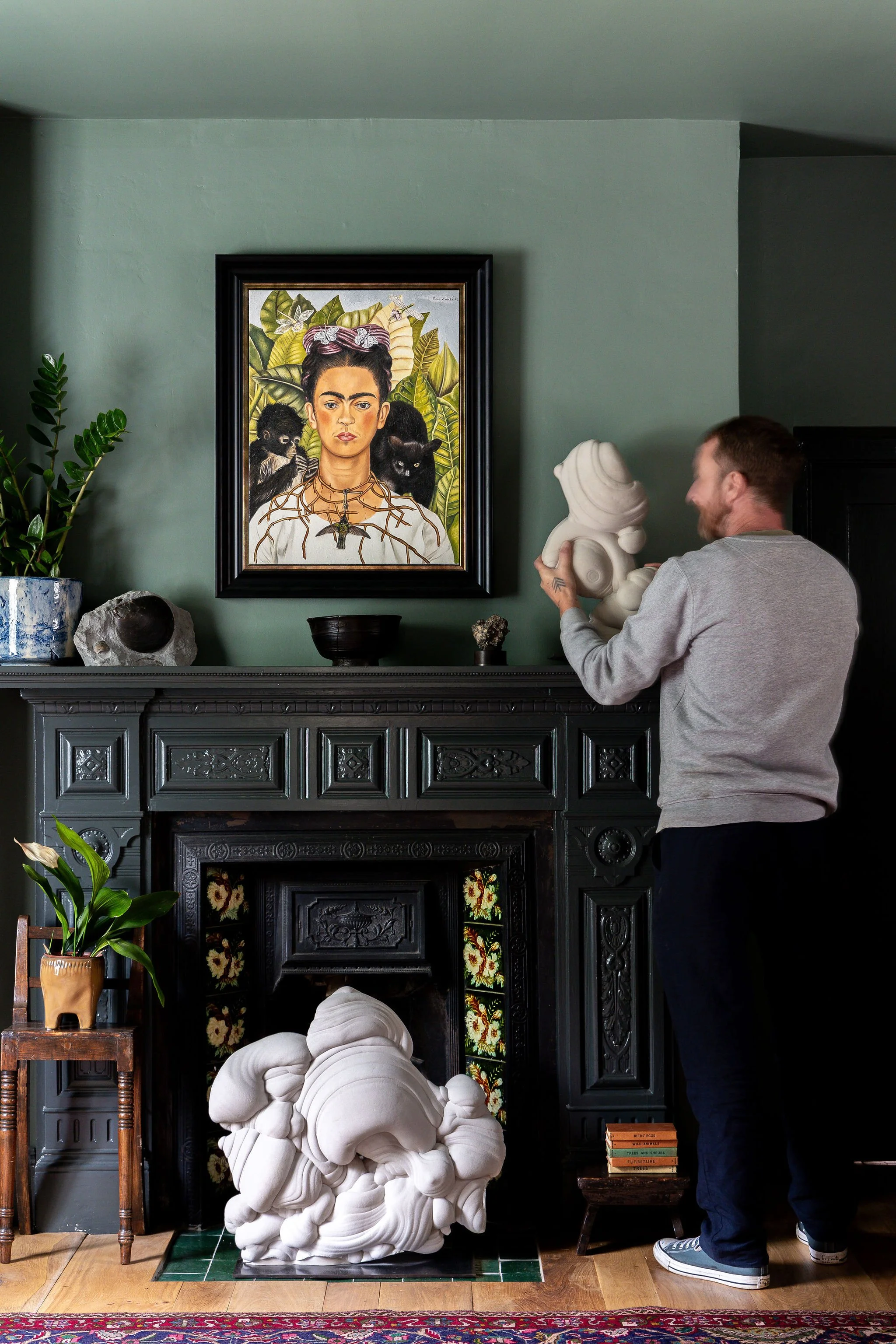 A man in a gray sweatshirt and blue pants arranging a white sculpture in a room with green walls, black ornate fireplace, colorful painting of Frida Kahlo with animals hanging above the fireplace, potted plants, and decorative objects.