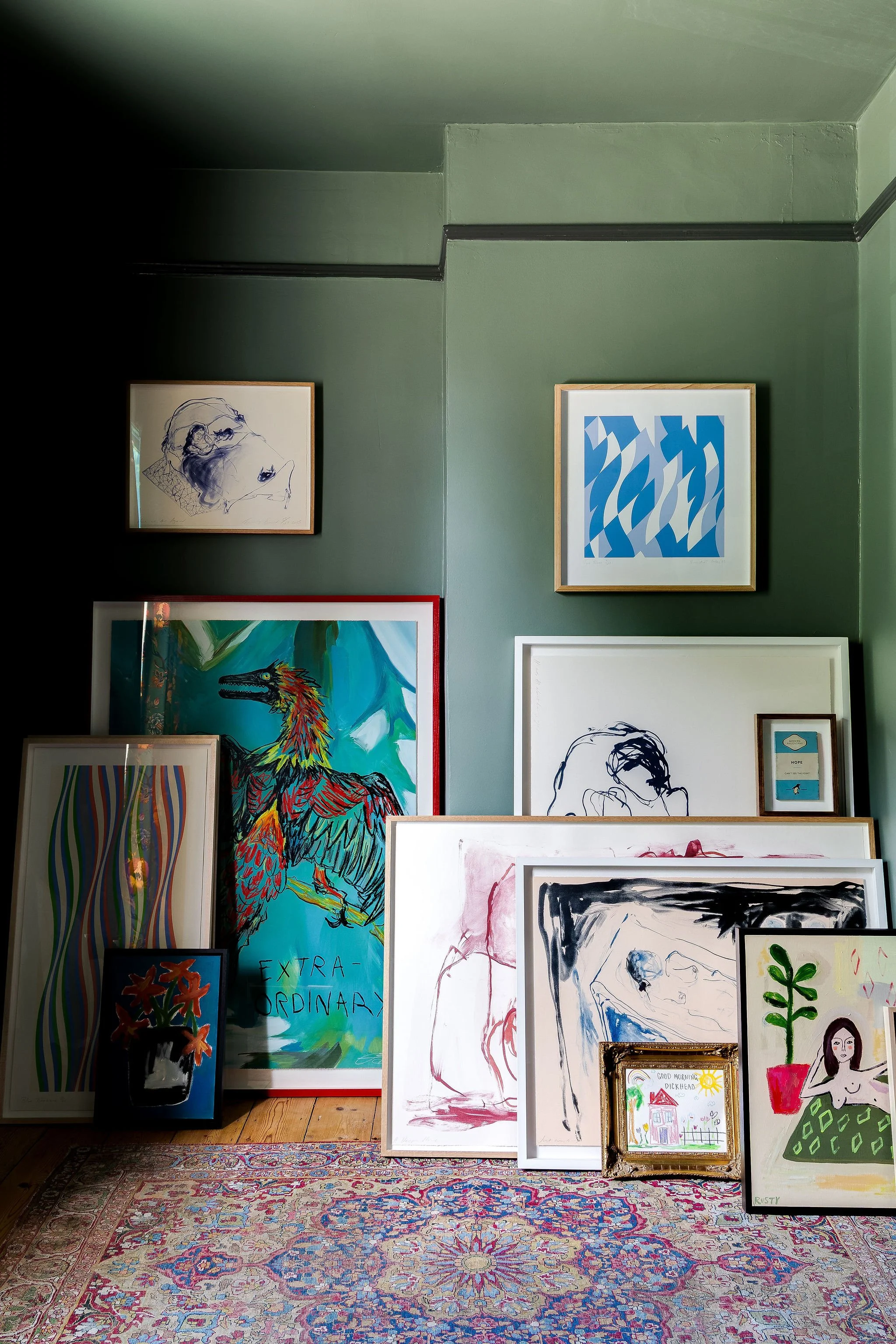 A collection of colorful paintings and drawings leaning against a green wall, with some framed and others unframed, on a patterned rug.