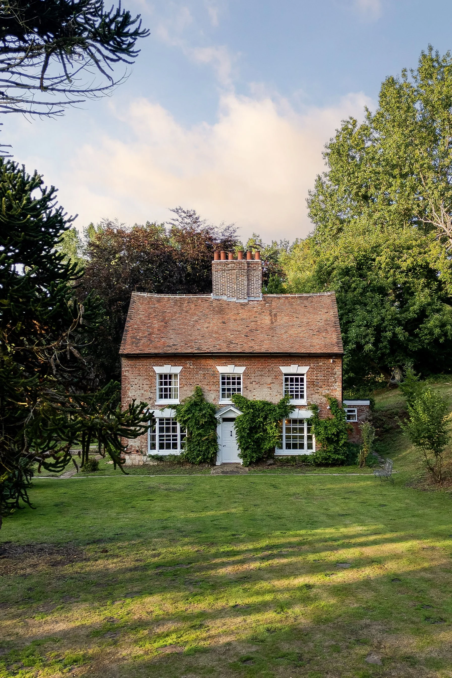 A brick house with white-framed windows and a white door, surrounded by green vines, situated on a grassy lawn with trees in the background under a partly cloudy sky.