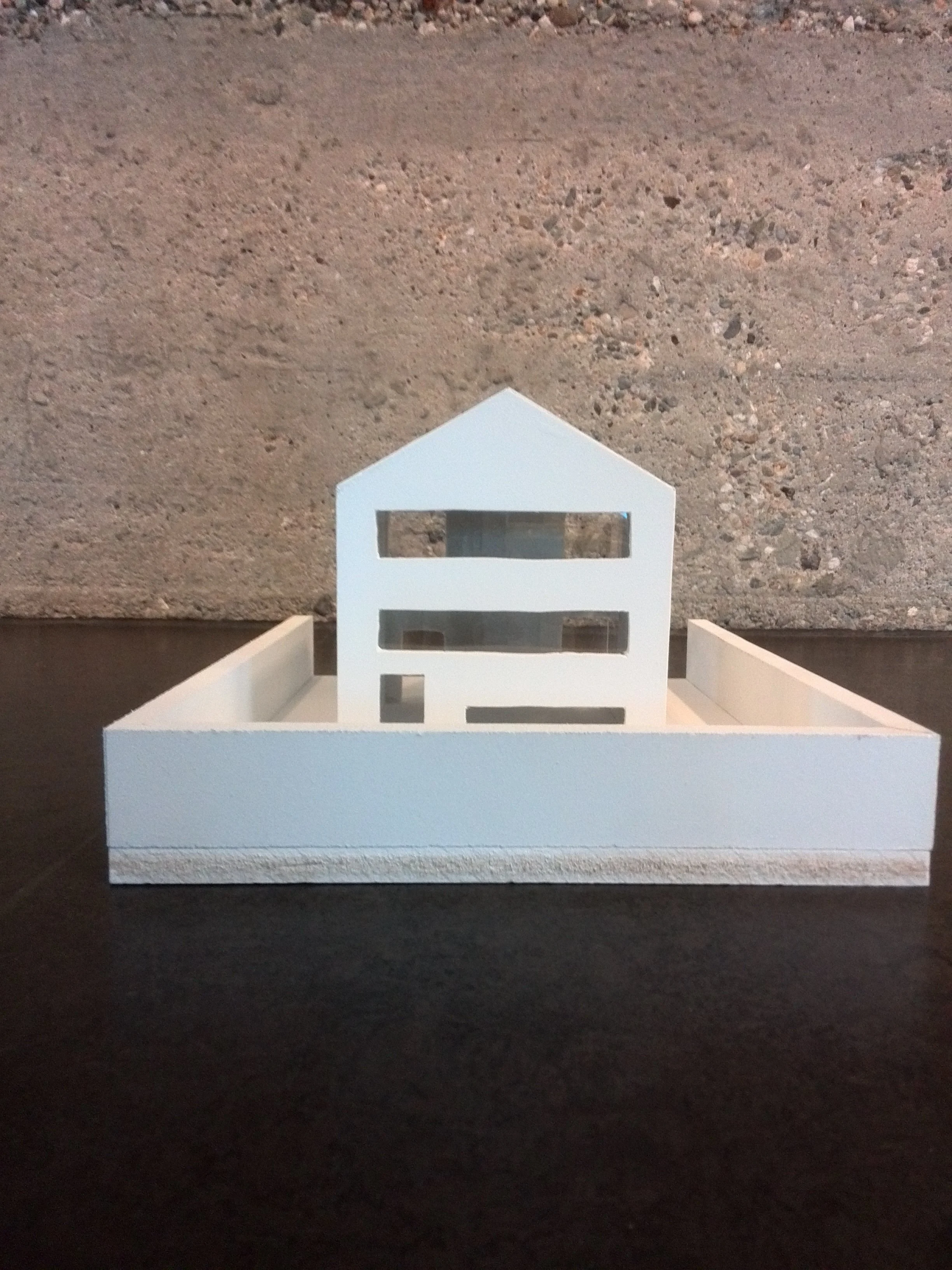 A small white model of a two-story house or building with a pitched roof, placed on a black surface against a gray, textured wall background.