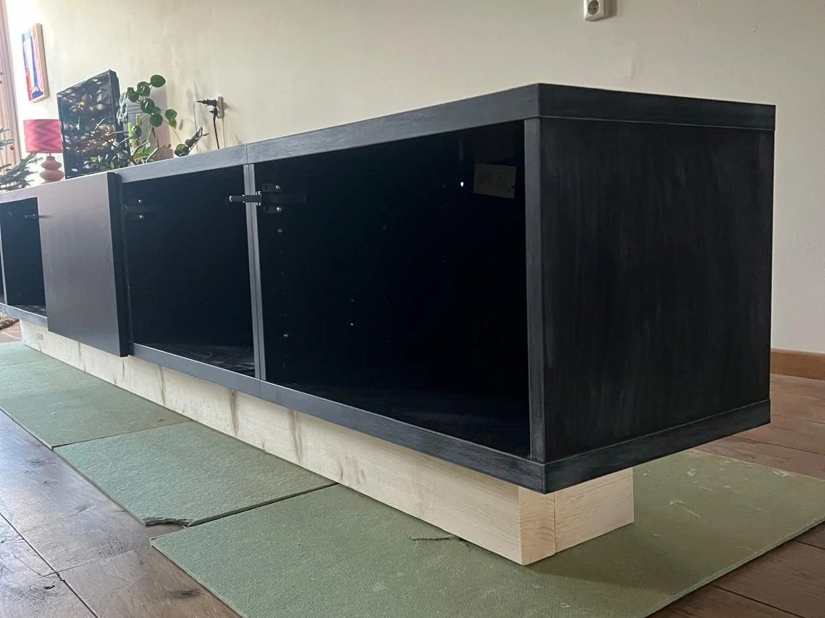 Een zwarte onderkast zonder deurtjes staat op een houten blokje, op een groene tapijt op een houten vloer in een kamer.