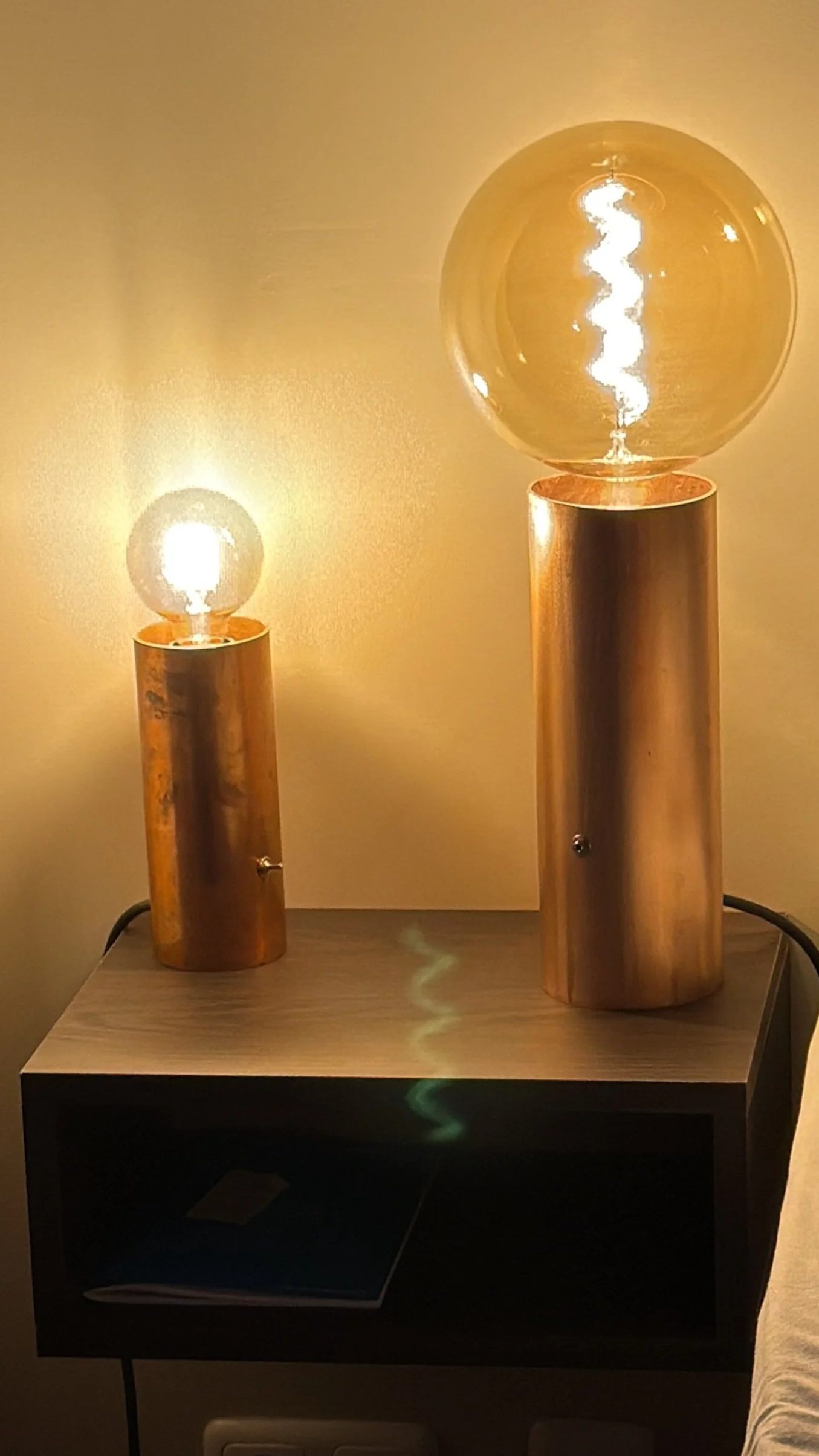Two decorative table lamps with clear glass bulbs and gold-colored bases, placed on a black nightstand against a yellow wall.