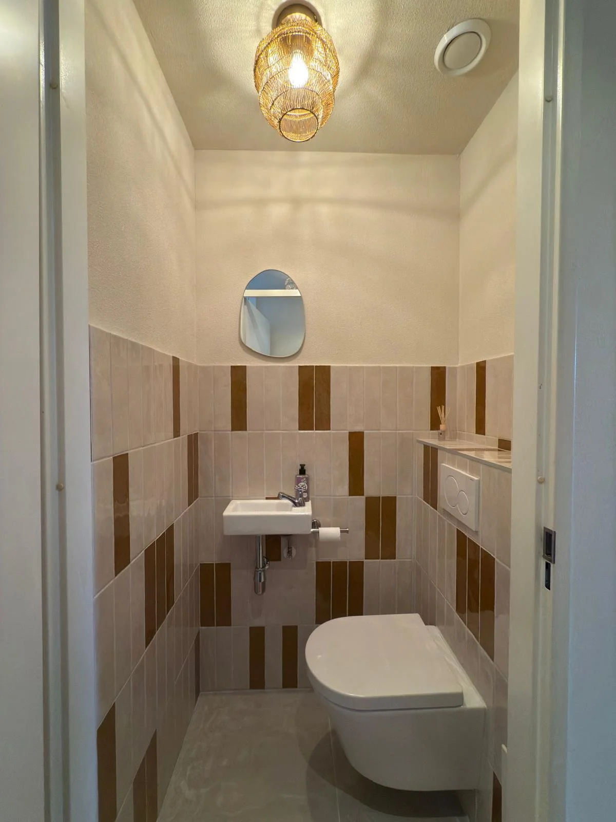 Small bathroom with beige and brown tiled walls, a white toilet, a tiny white sink, a small oval mirror, a woven pendant light, and a small shelf with reed diffusers.