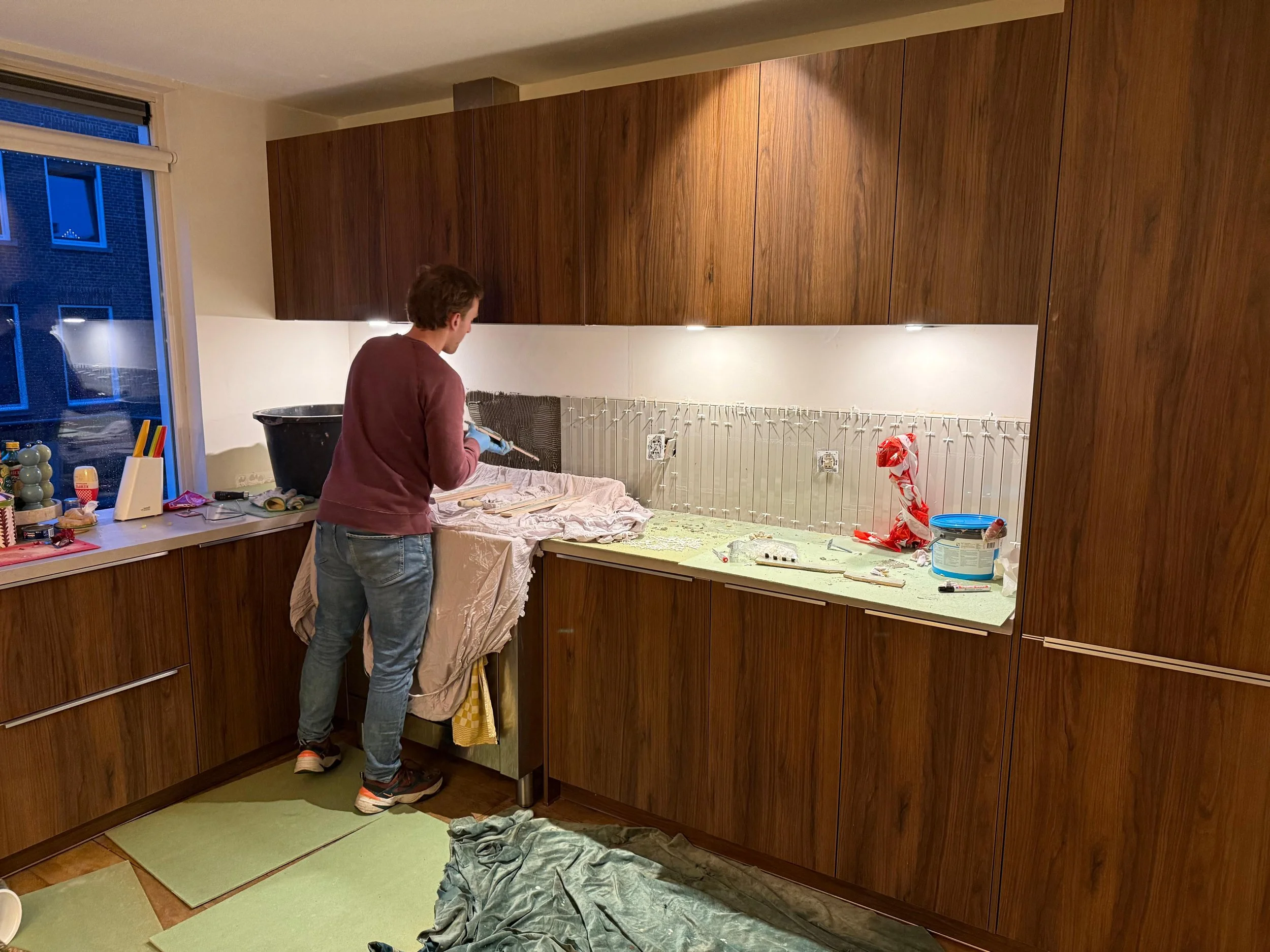 Een persoon die verf spuit op de muur van een keuken met houten kasten, terwijl hij op een kruk staat. Er zijn dozen en verfbenodigdheden op het aanrecht en op de vloer ligt een stuk doek om de vloer te beschermen.