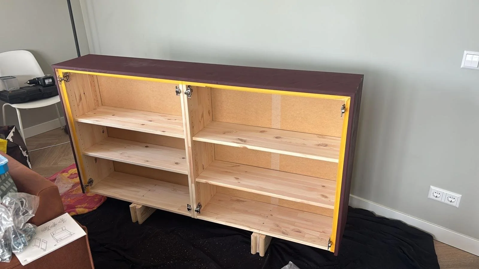 Onbehandelde houten kast met drie planken en twee deuren, geplaatst in een kamer met witte muur.