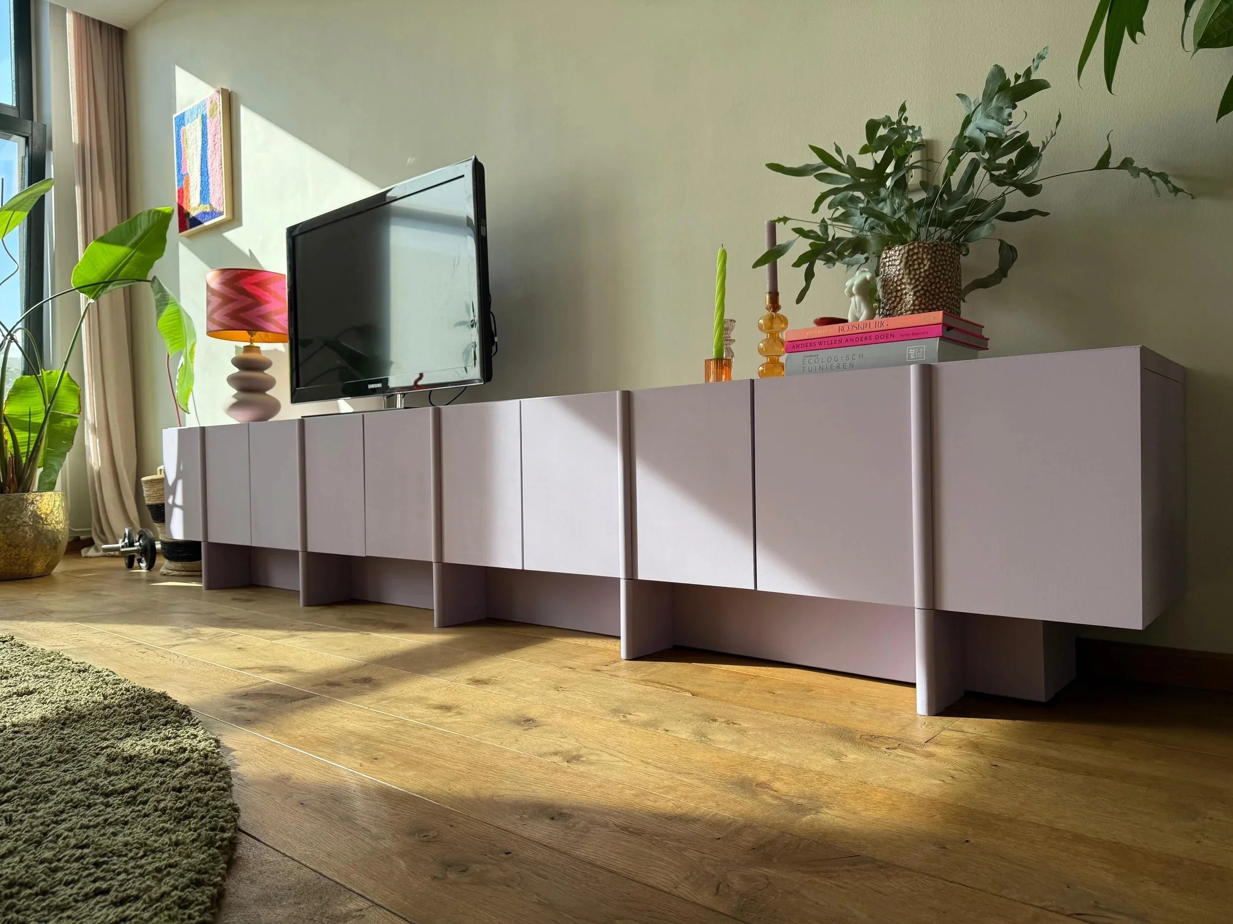 Een woonkamer met een tv op een wit meubel, omringd door planten en decoratieve items, met houten vloer en grote ramen voor natuurlijk licht.