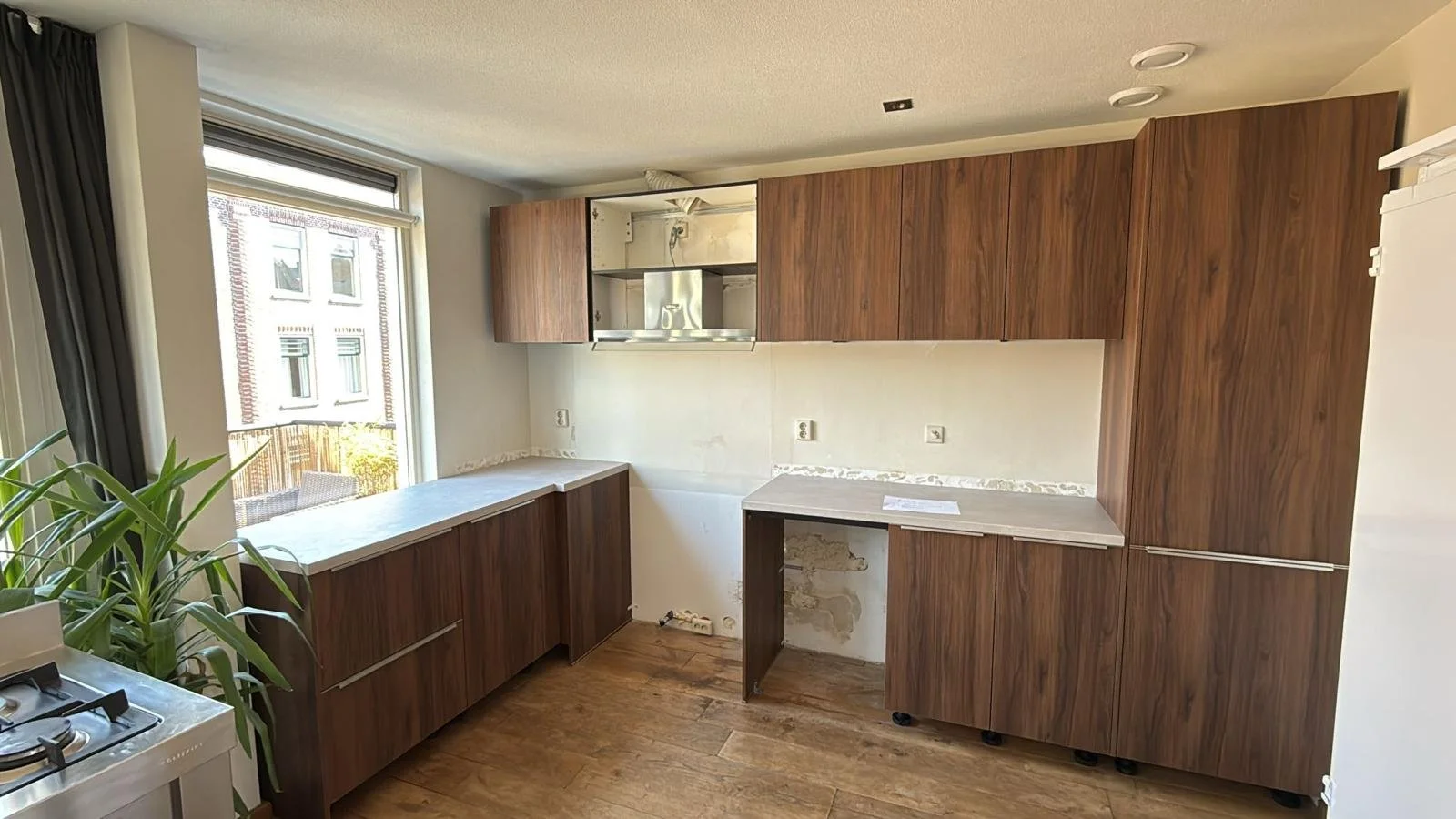 Een keuken in opknapbeurt met houten kasten, een groot raam, en een gasfornuis op de linkerkant.