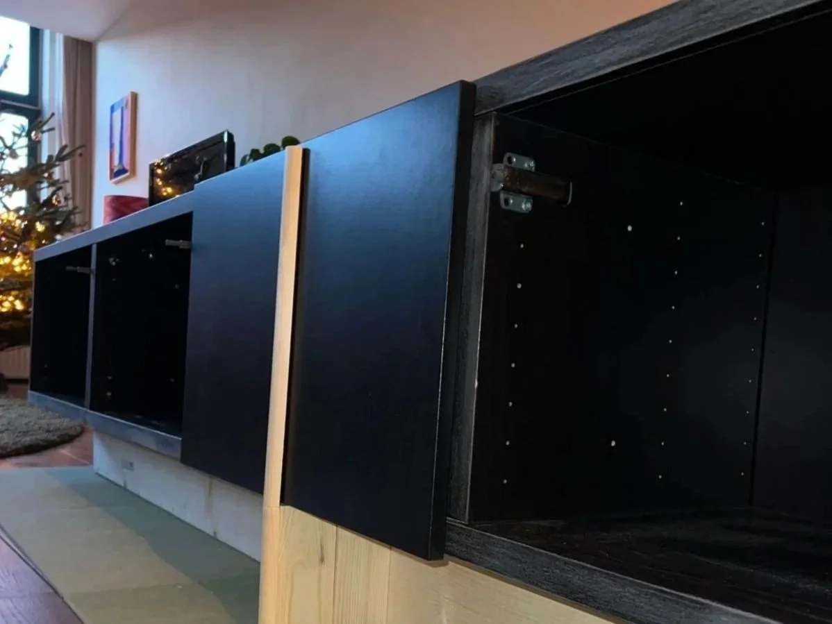 Een zwarte houten kast onder een televisie in een woonkamer, met een kerstboom zichtbaar op de achtergrond.