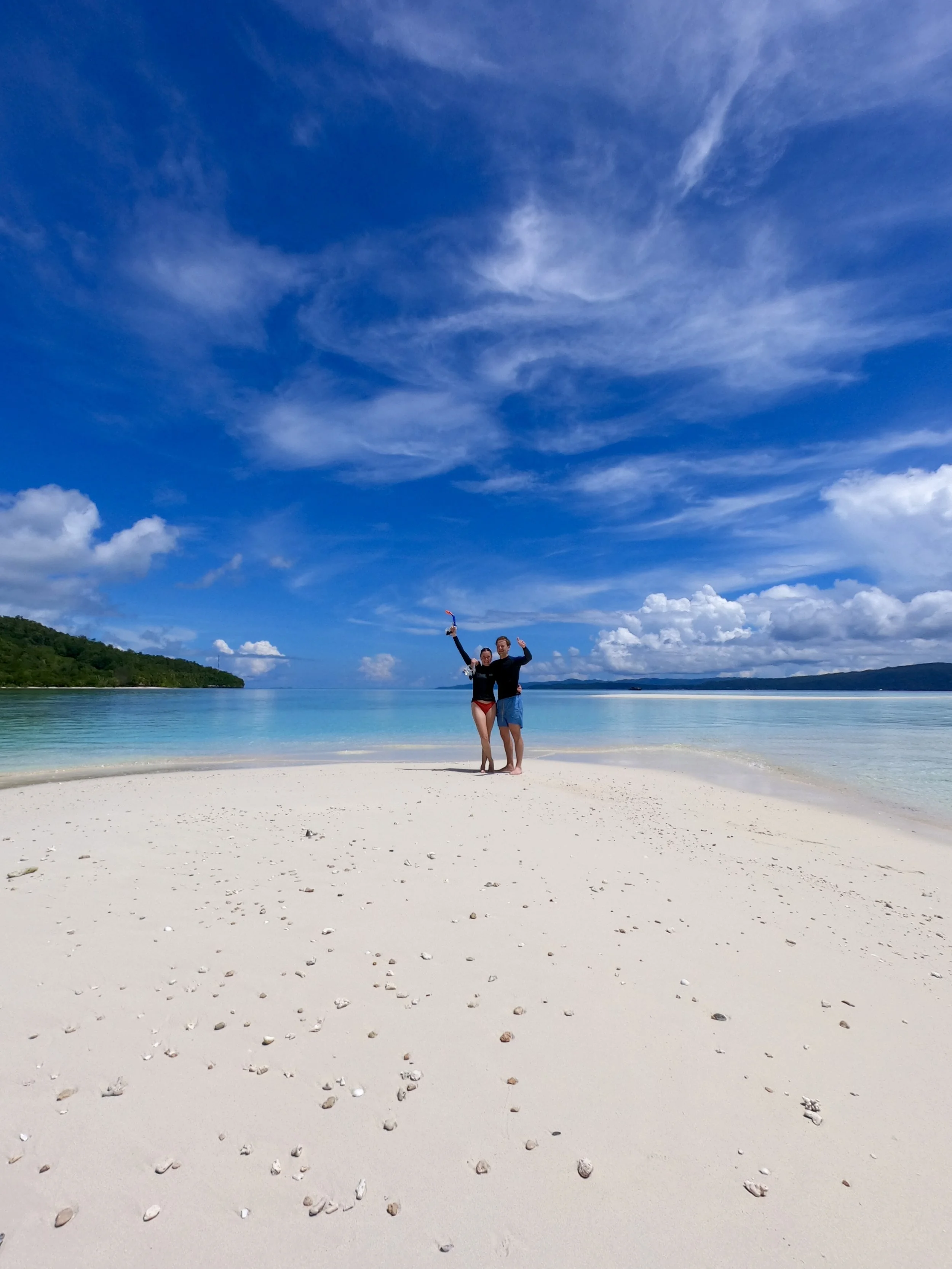 Two people standing on a sandy beach with clear water, a lush hillside on the left, and a partly cloudy blue sky above.