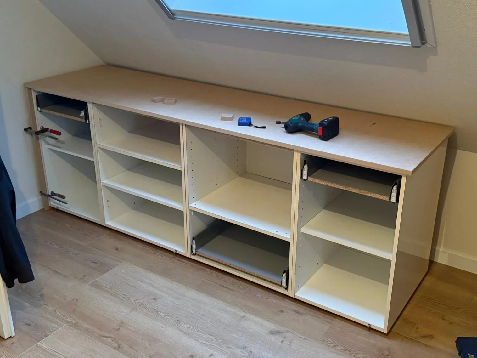 Onbeveiligde houten kast met open planken onder schuine dakramen, gereedschap en bouwmaterialen op de bovenkant.