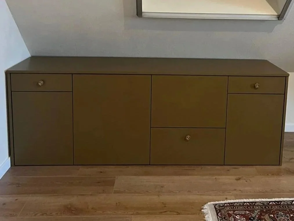 Een groene houten buffetkast met vier laden, geplaatst tegen een muur met houten vloer en een vloerkleed links.