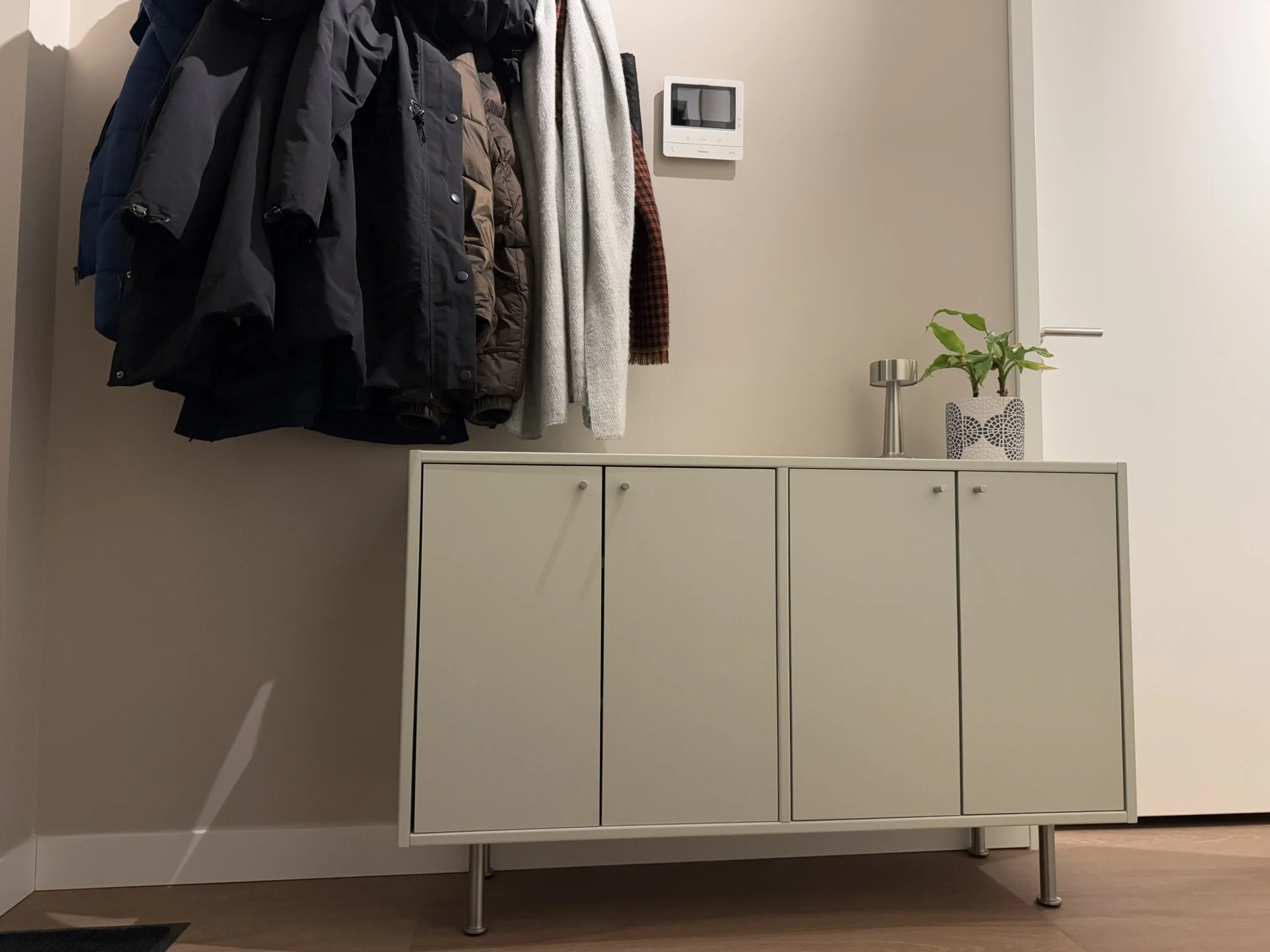 Coat rack with jackets and scarves above a white cabinet with a potted plant and a small lamp on top, adjacent to a beige wall with a thermostat.
