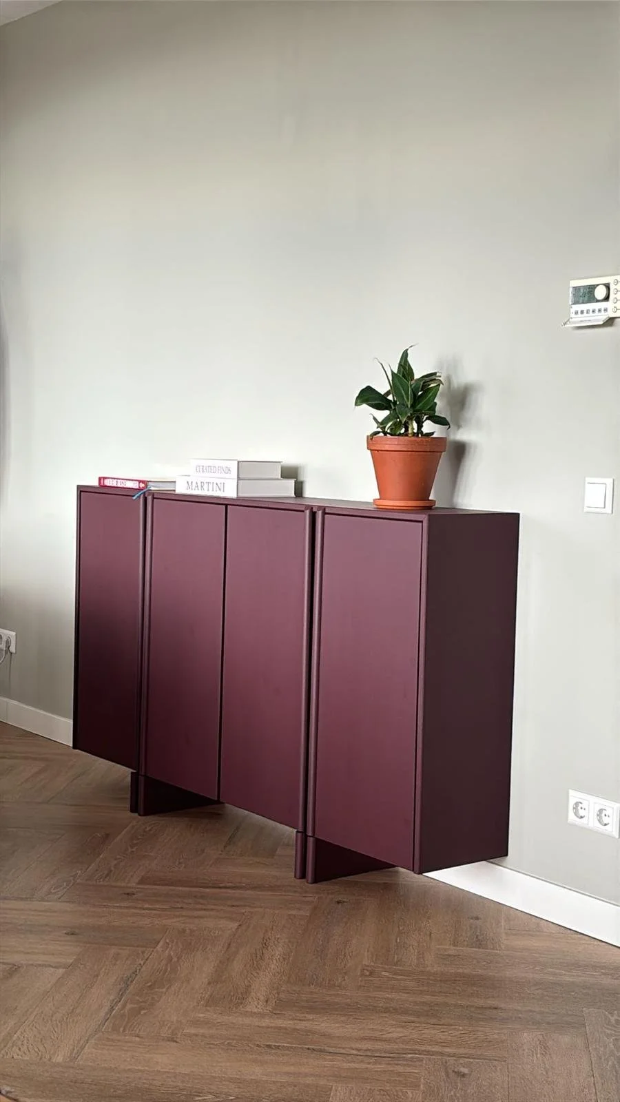 Een muur met een lage kast met een paar boeken en een plant in een pot op de kast, en een vloer met houtpatroon.