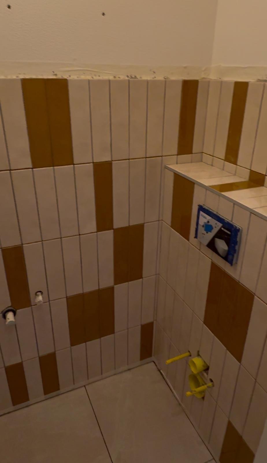 An unfinished bathroom wall with beige and brown tiles, plumbing pipes, and an electrical outlet box.