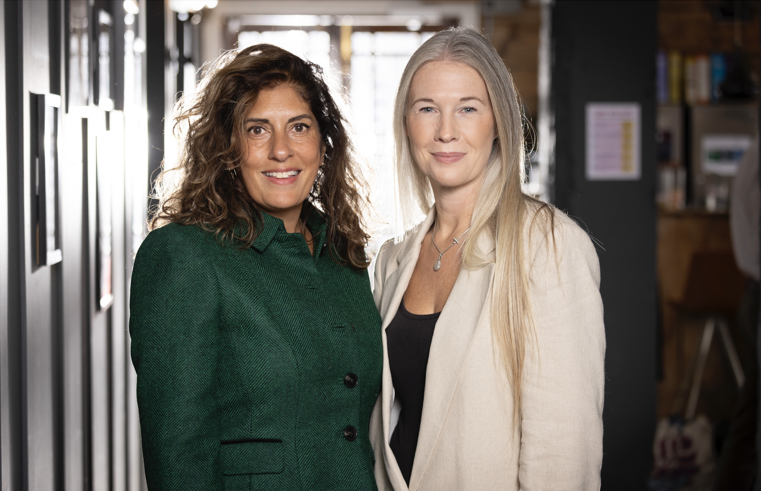 Two women standing close together indoors, smiling at the camera, with a blurred background.