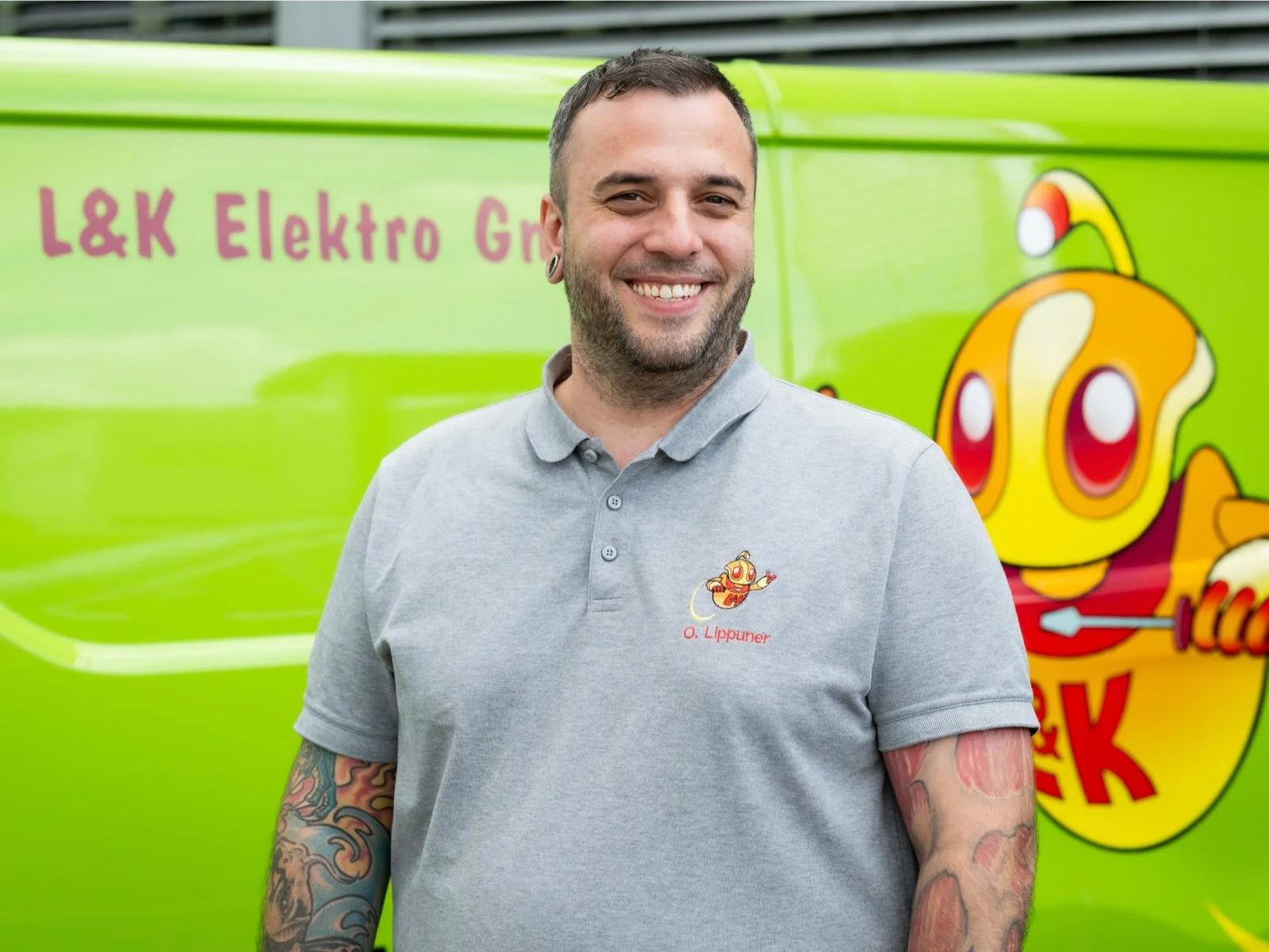 Ein lachender Mann in einem grauen Polo-Shirt mit einem Cartoon-Logo auf der Brust, vor einem grünen Fahrzeug mit einem bunten Cartoon-Charakter und textartigen Elementen.